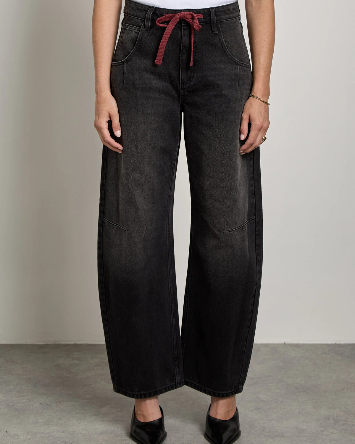 A woman stands in a studio wearing a white sleeveless top and wide-leg black jeans with a red drawstring at the waist, paired with black pointed shoes.