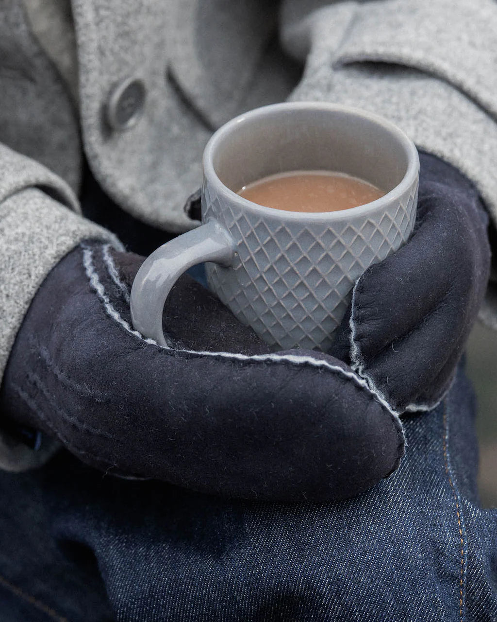 Person wearing gloves holding a white mug with a warm beverage, wearing a coat.