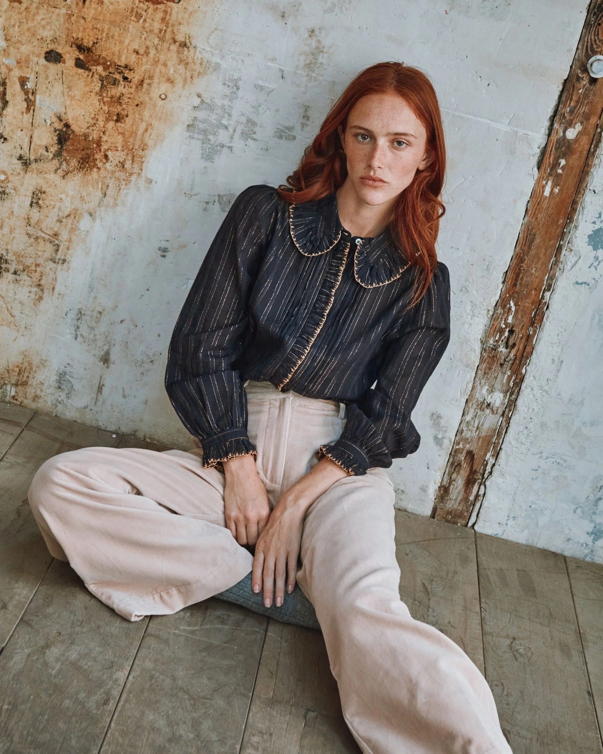 Woman sitting on the floor wearing a dark blouse and light pants against a rustic wall.