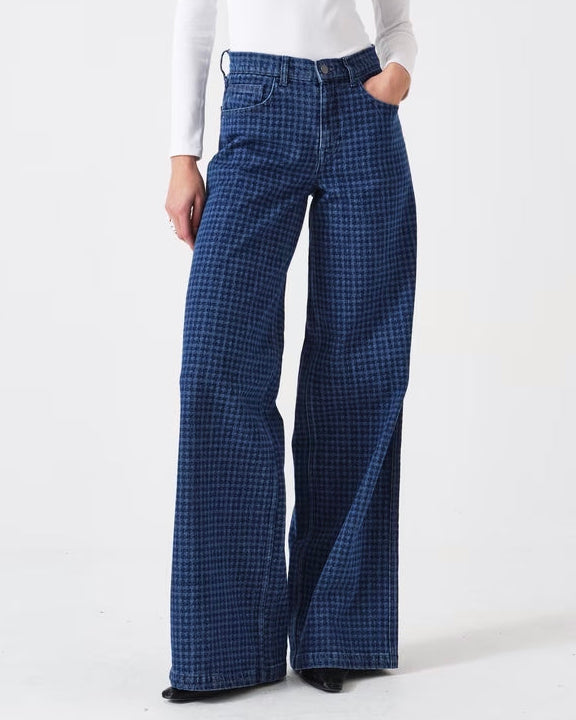 A model stands against a plain white studio background wearing a white long-sleeve henley top and dark blue wide-leg plaid trousers.
