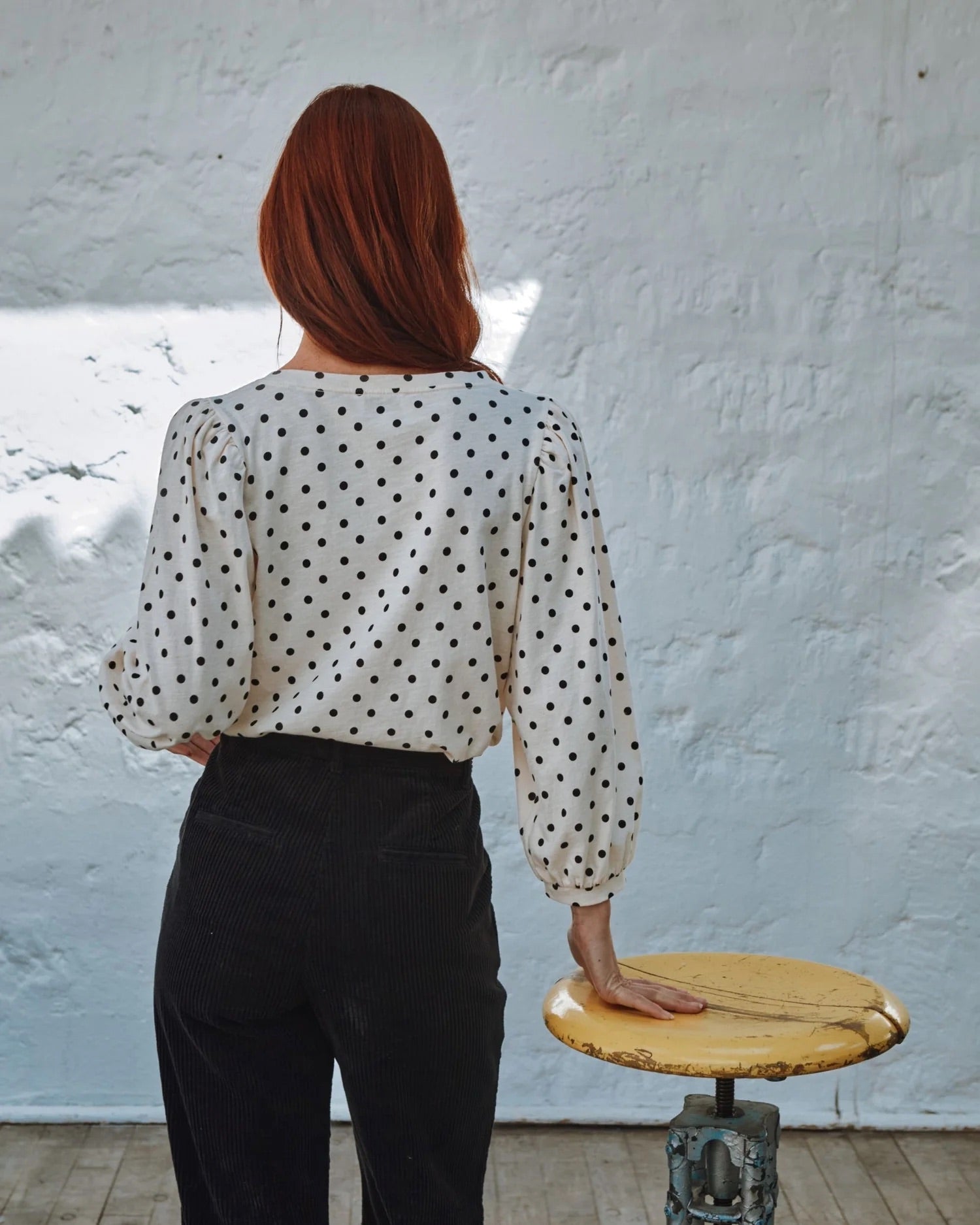 Person wearing a polka dot blouse and black pants standing next to a wooden stool against a textured white wall.