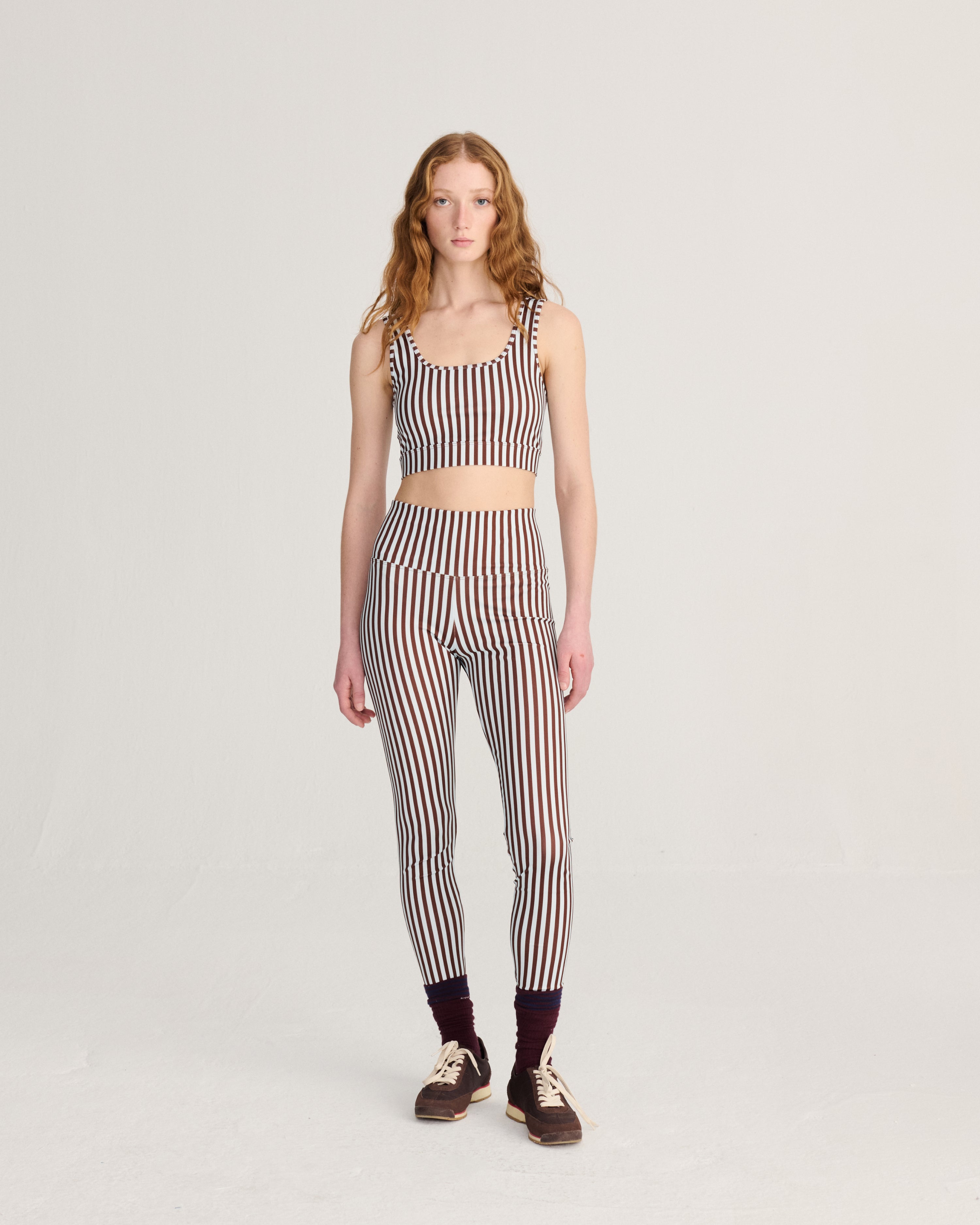 A model stands against a light gray studio background wearing a matching two-piece set with vertical stripes in dark brown, beige, and white. The set includes a sleeveless crop top and high-waisted leggings, paired with dark socks and brown sneakers.