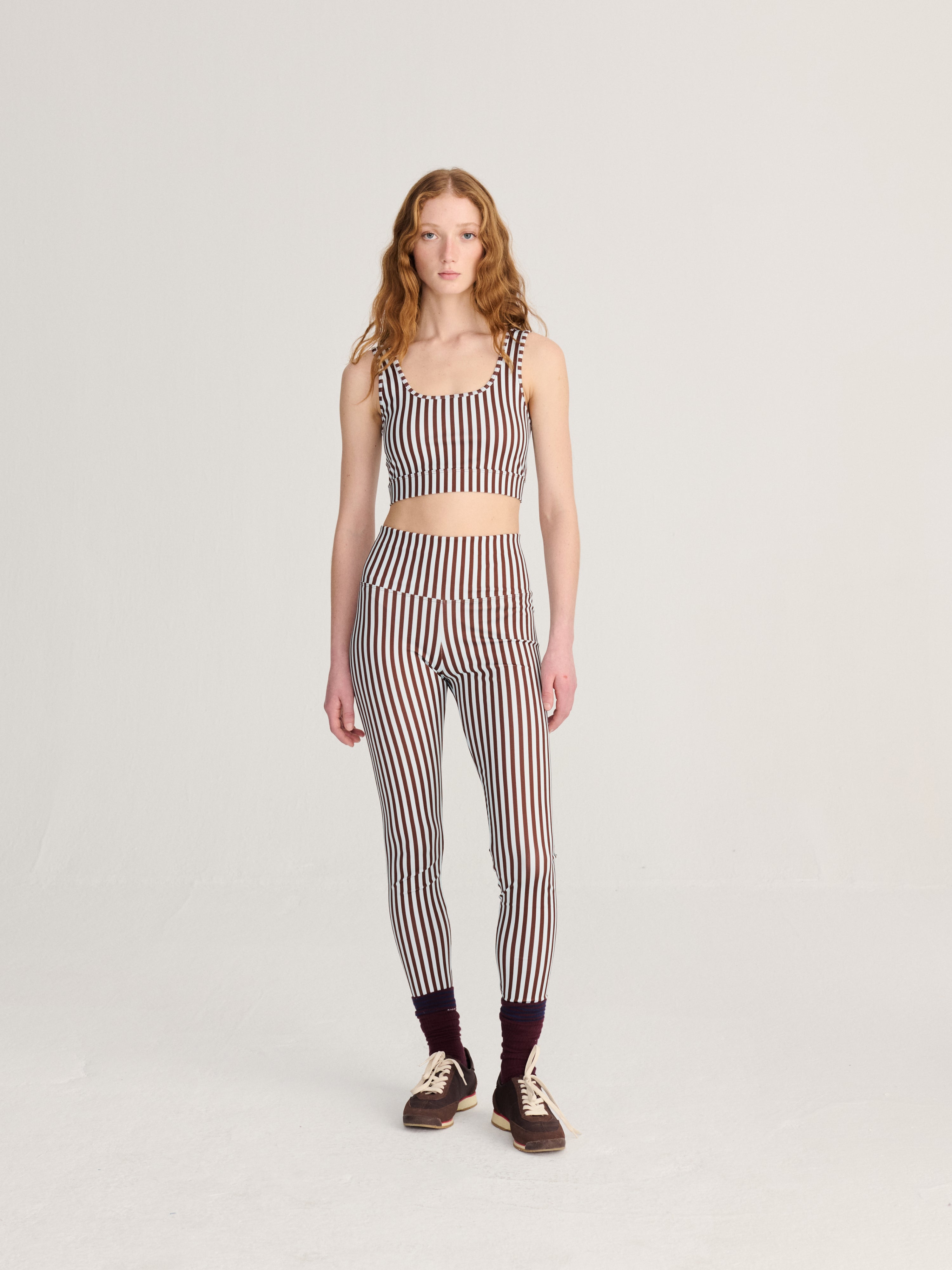 A model stands against a light gray studio background wearing a matching two-piece set with vertical stripes in dark brown, beige, and white. The set includes a sleeveless crop top and high-waisted leggings, paired with dark socks and brown sneakers.