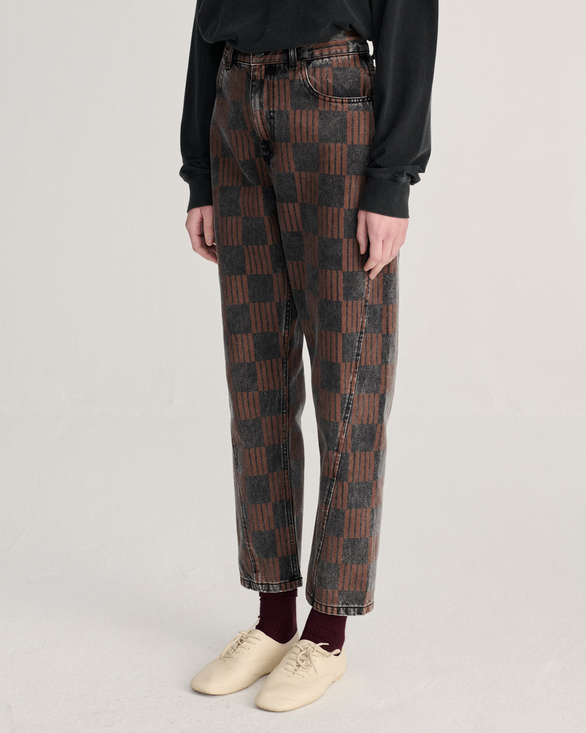A person wearing patterned brown and black checkered jeans, a black long-sleeve top, burgundy socks, and cream-colored sneakers, standing against a light gray studio background.