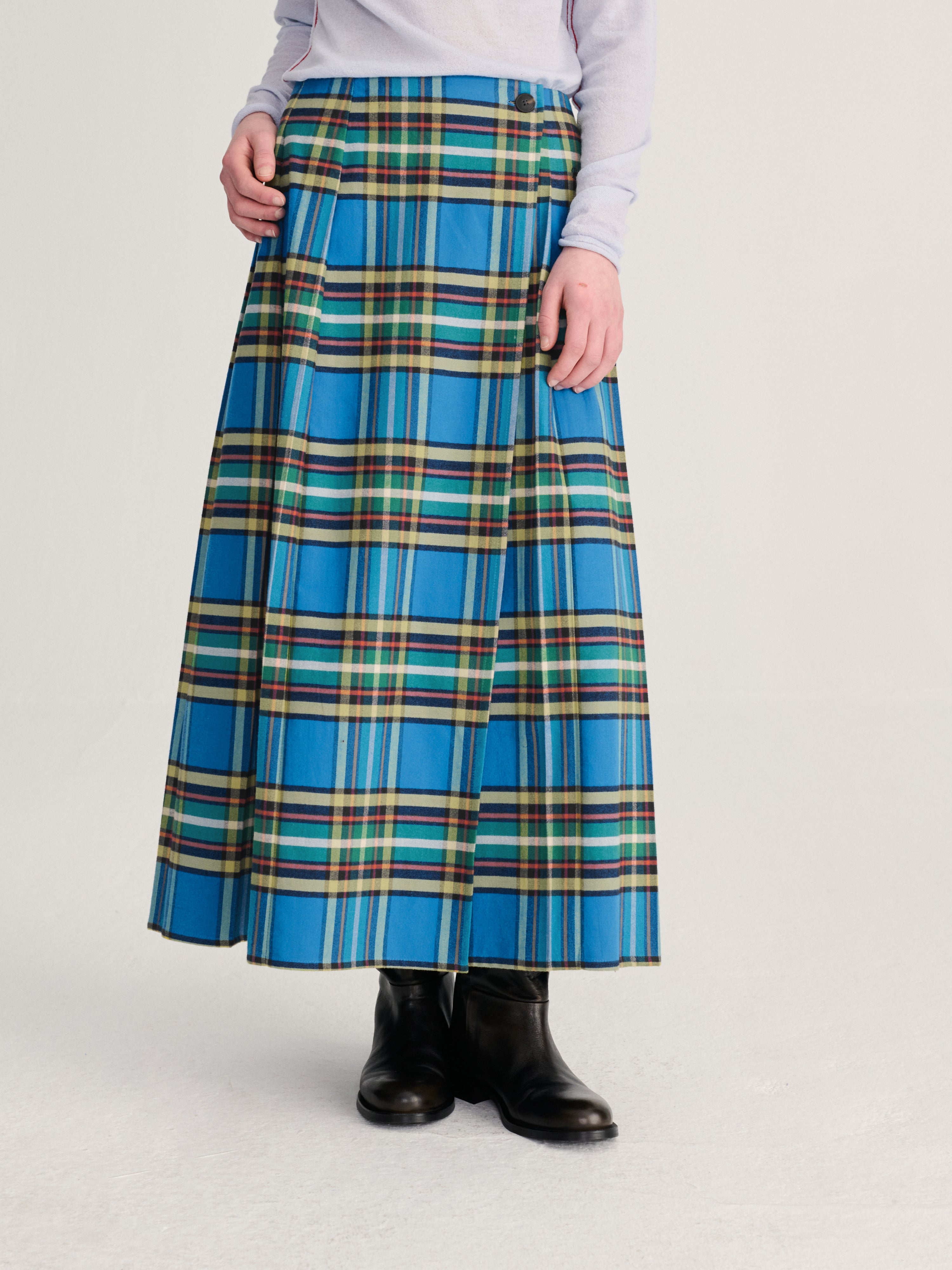 A model wearing a high-waisted, pleated A-line skirt in a vibrant blue tartan plaid pattern with green, yellow, and white stripes, styled with a light gray top and black leather ankle boots against a light gray studio background.