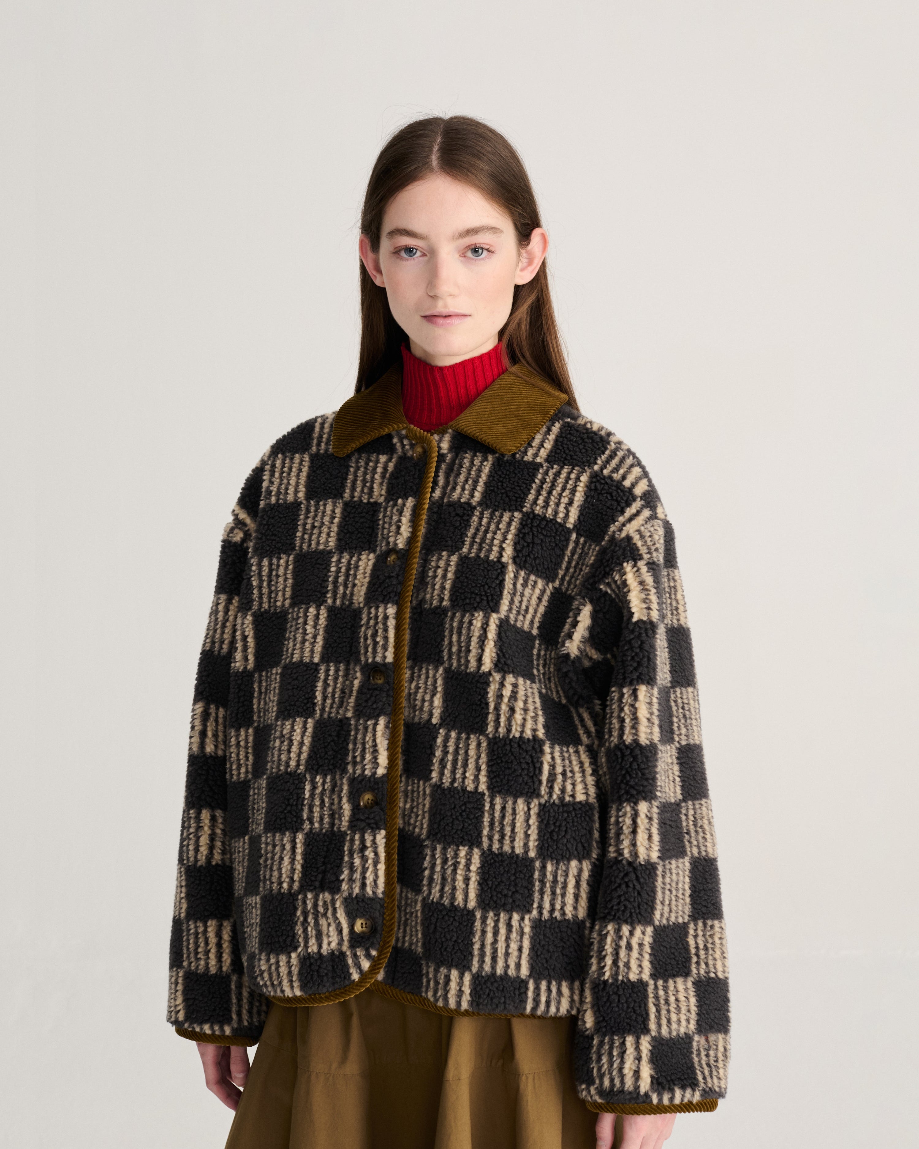 A model wearing a black and beige checkered textured jacket with a brown corduroy collar and brown buttons, layered over a red turtleneck and brown skirt, against a light gray studio background.