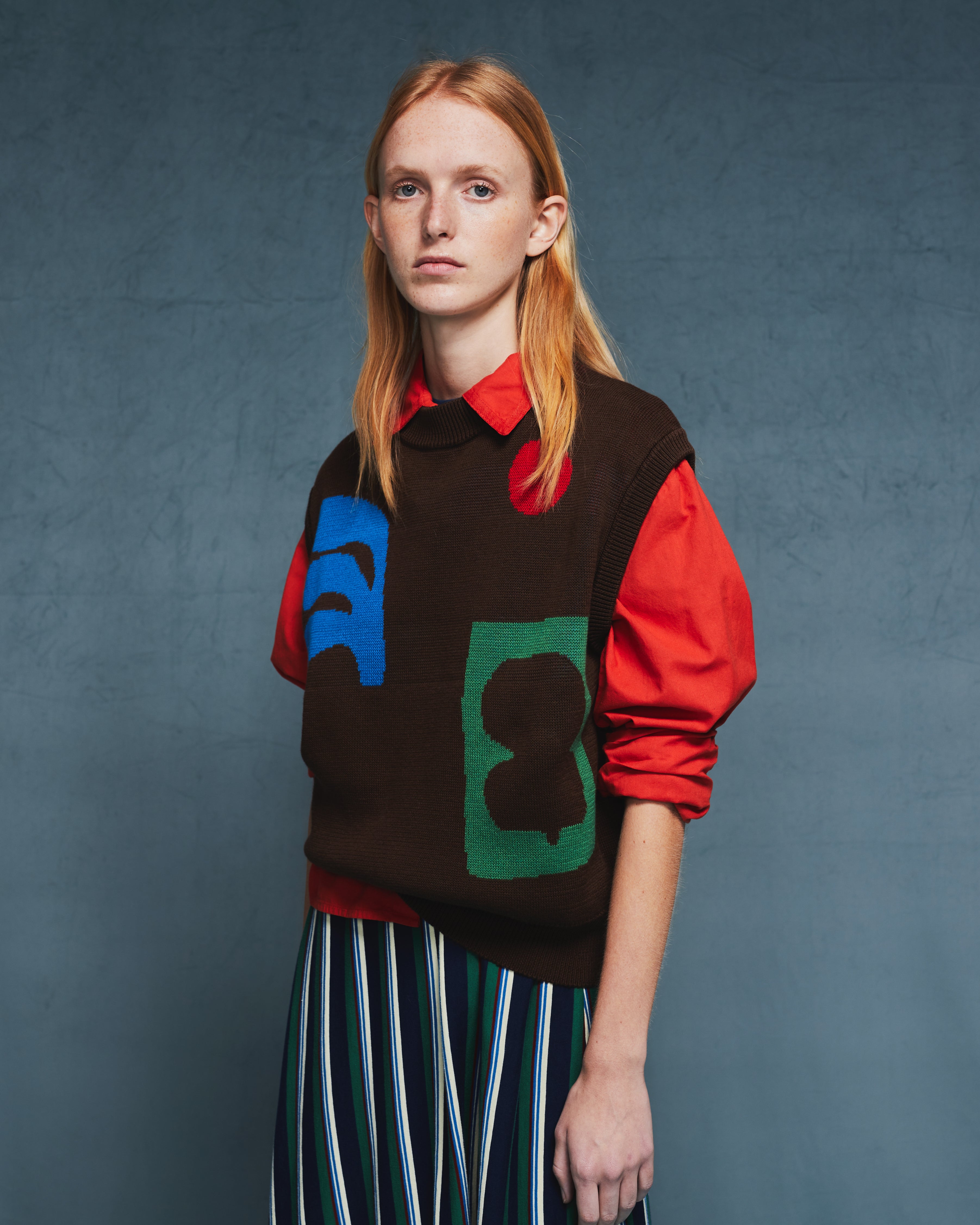 A model wearing a brown knit sweater vest with colorful abstract patches over a red long-sleeve shirt and a green, white, and black striped skirt, set against a textured dark blue-gray studio background.