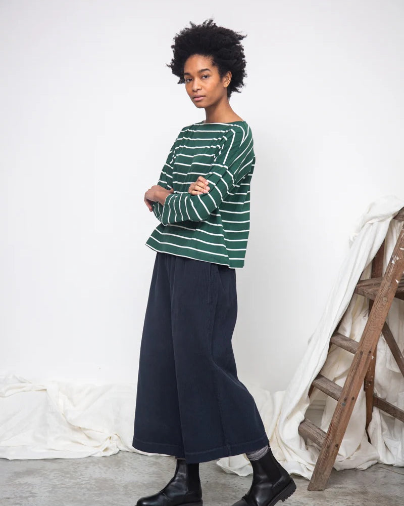 Person wearing a green striped sweater and dark blue skirt standing against a white wall with a wooden ladder.