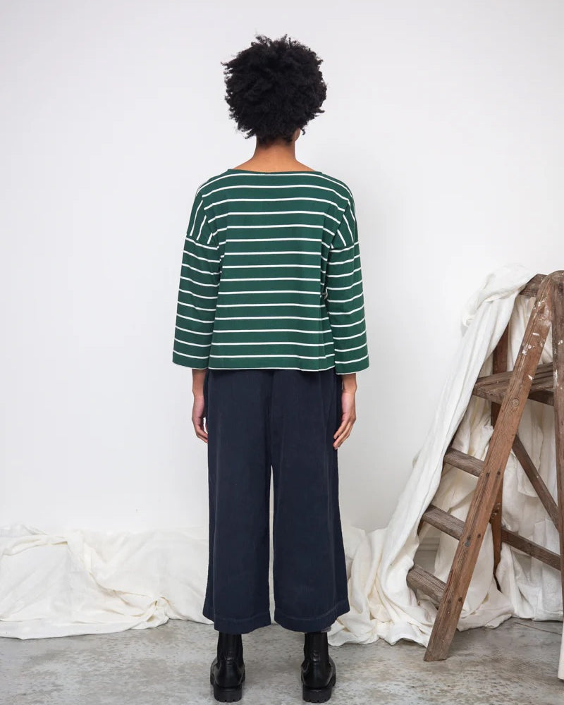 Person wearing a green and white striped sweater and dark pants standing against a white wall with a wooden ladder.