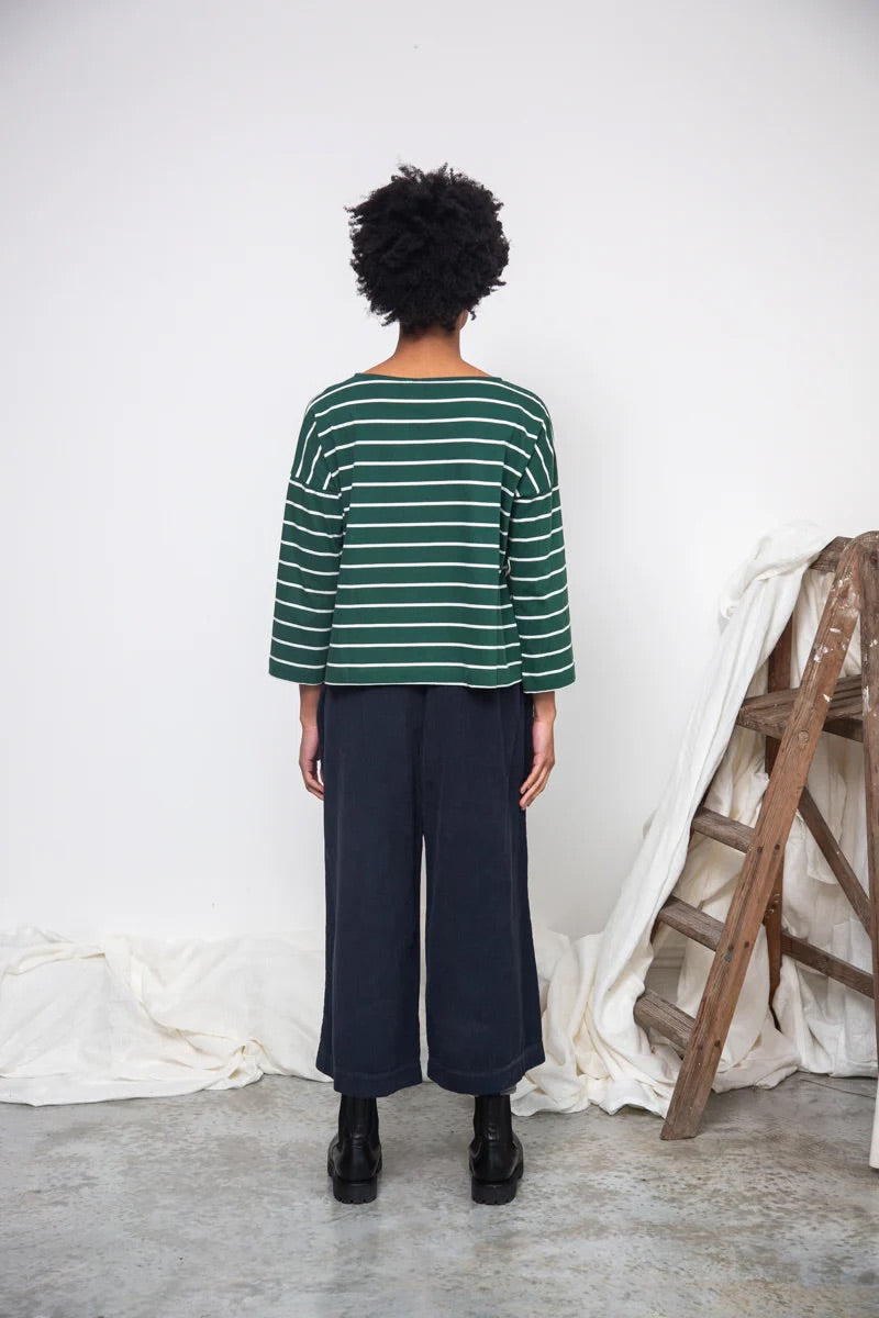 Person wearing a green and white striped sweater and dark pants standing against a white wall with a wooden ladder.