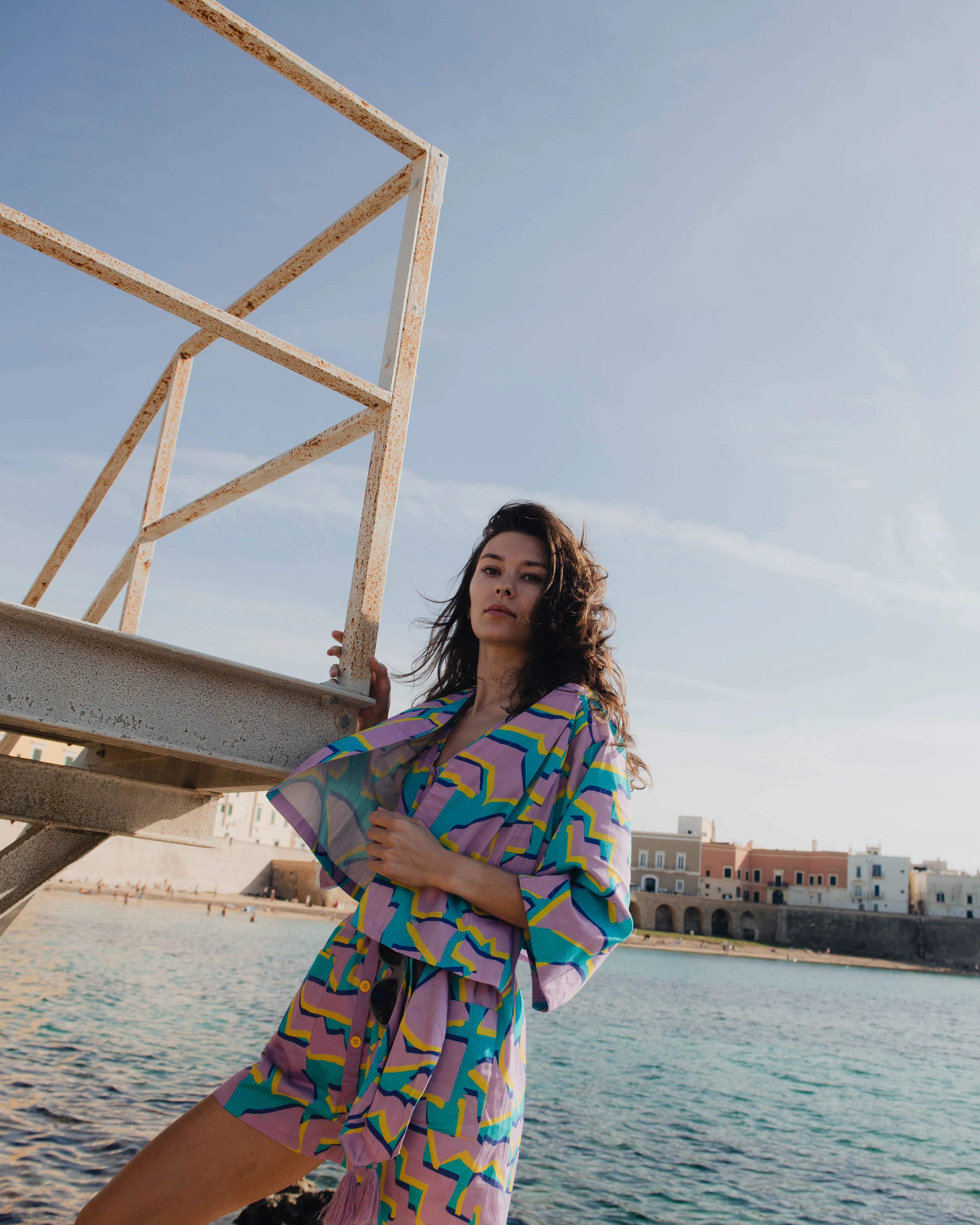 Woman in colorful outfit standing by a waterfront with buildings in the background.