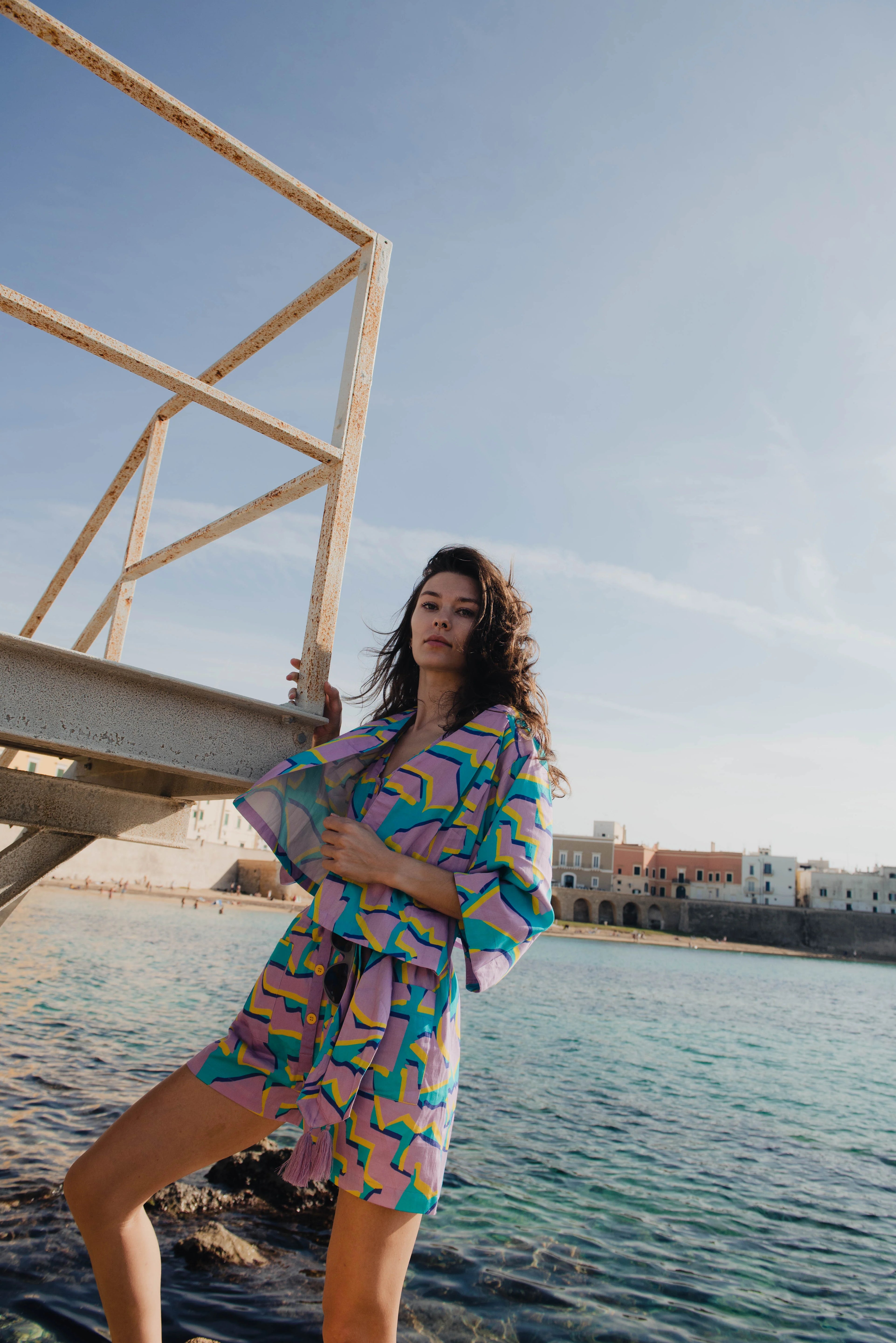 Woman in colorful outfit standing by a waterfront with buildings in the background.