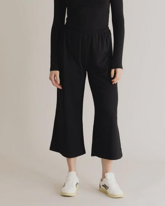 A model wearing black lounge pants with an elastic waistband and a relaxed fit. The pants are made from organic cotton and elastane, and the model is standing with one foot on a white shoe.