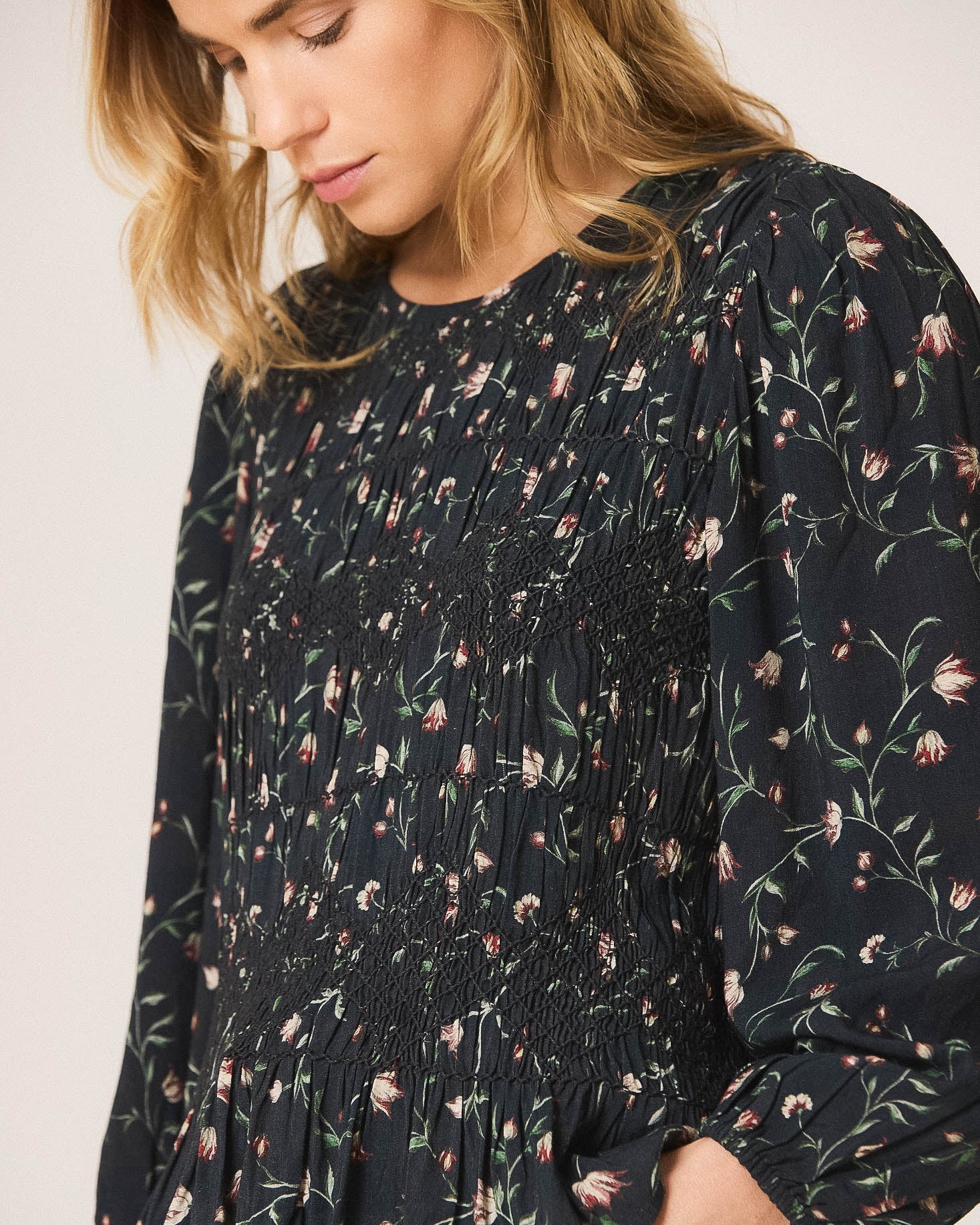 A woman wearing a dark navy floral long-sleeve blouse with a smocked bodice and peplum hem, paired with dark jeans against a plain light background.