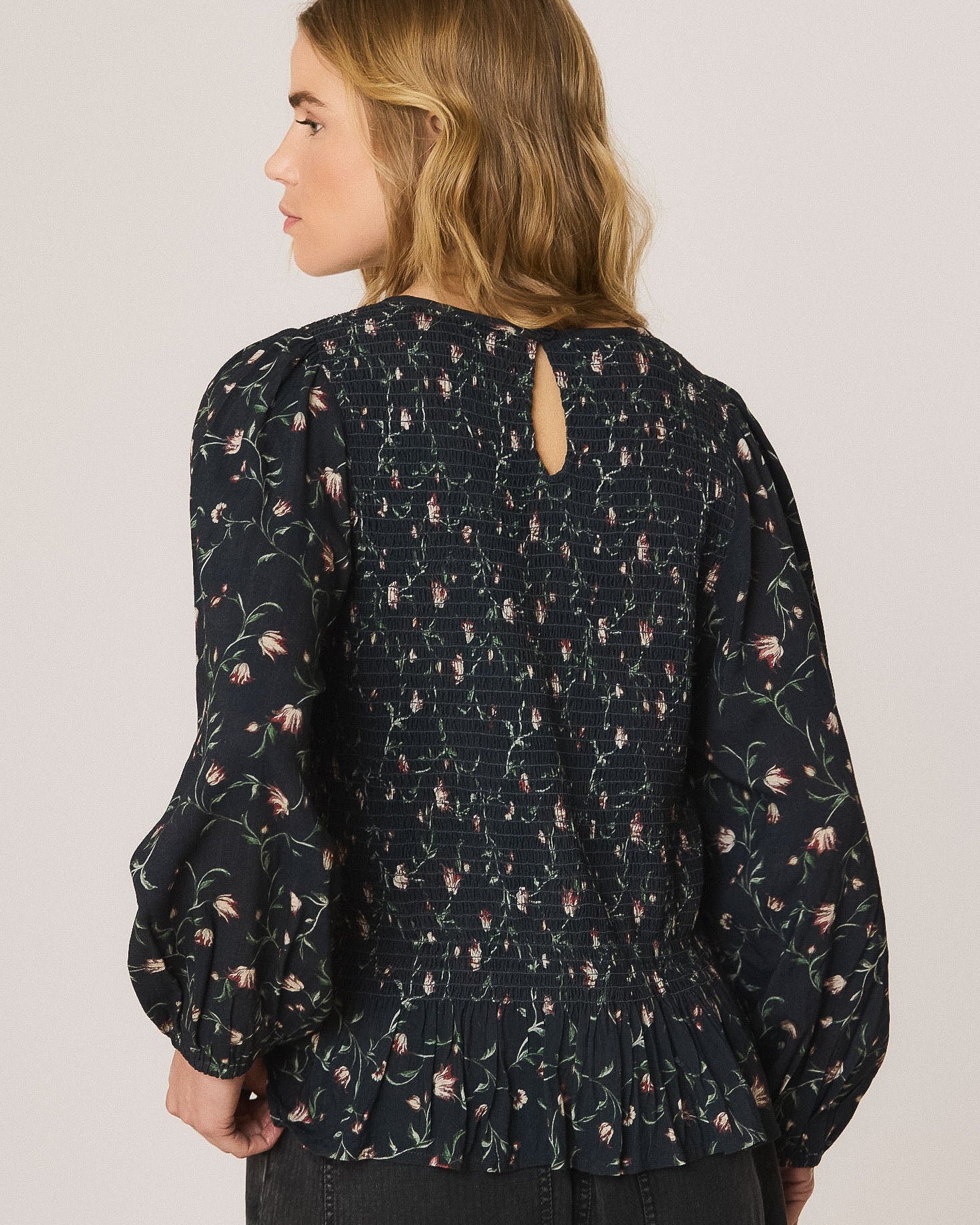 A woman shown from the back wearing a dark navy floral long-sleeve blouse with puff sleeves and a small keyhole cutout at the neckline, paired with dark jeans against a light gray studio background.