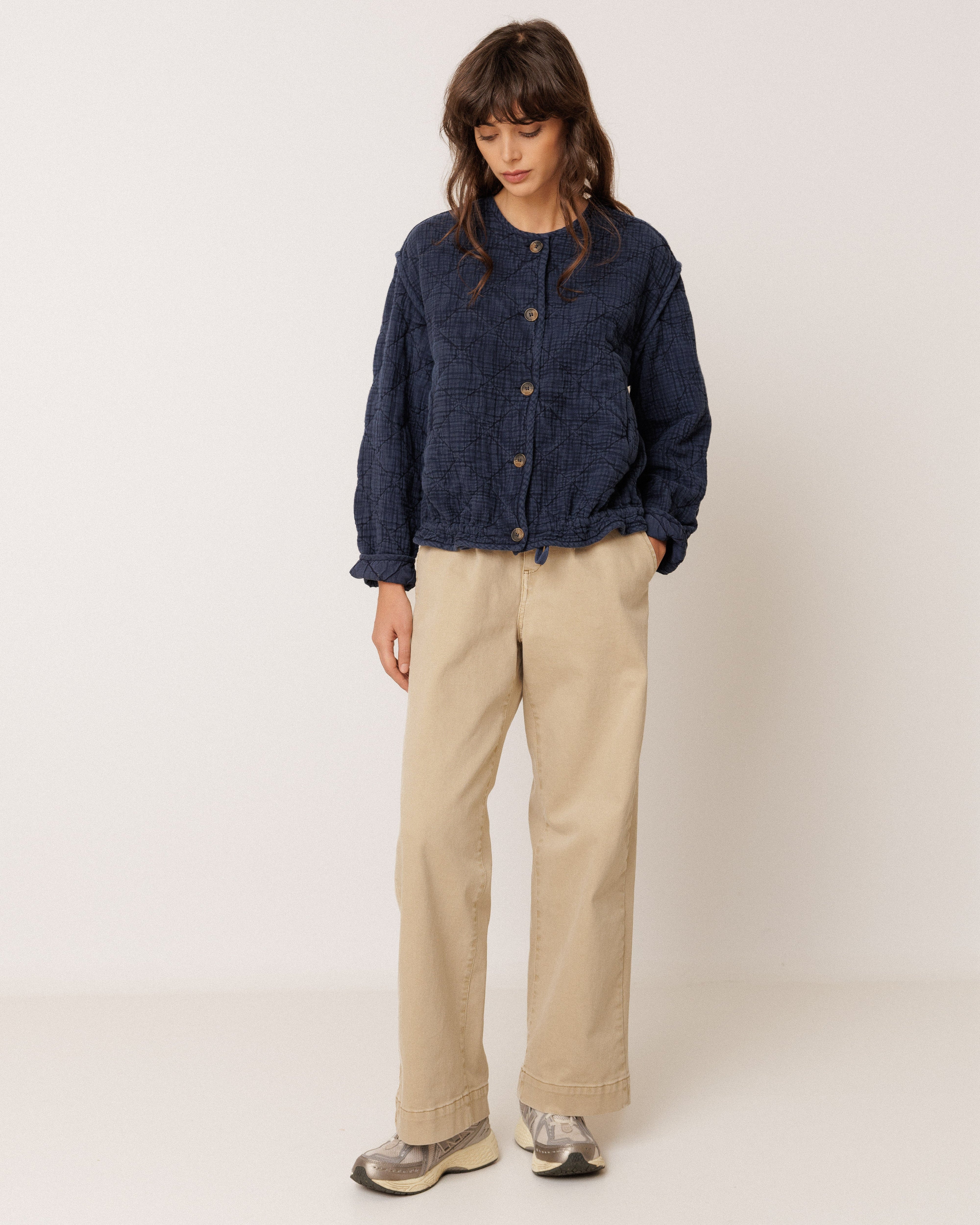 A model stands against a plain off-white studio background wearing a dark navy blue cropped button-up jacket, beige wide-leg trousers, and gray sneakers.
