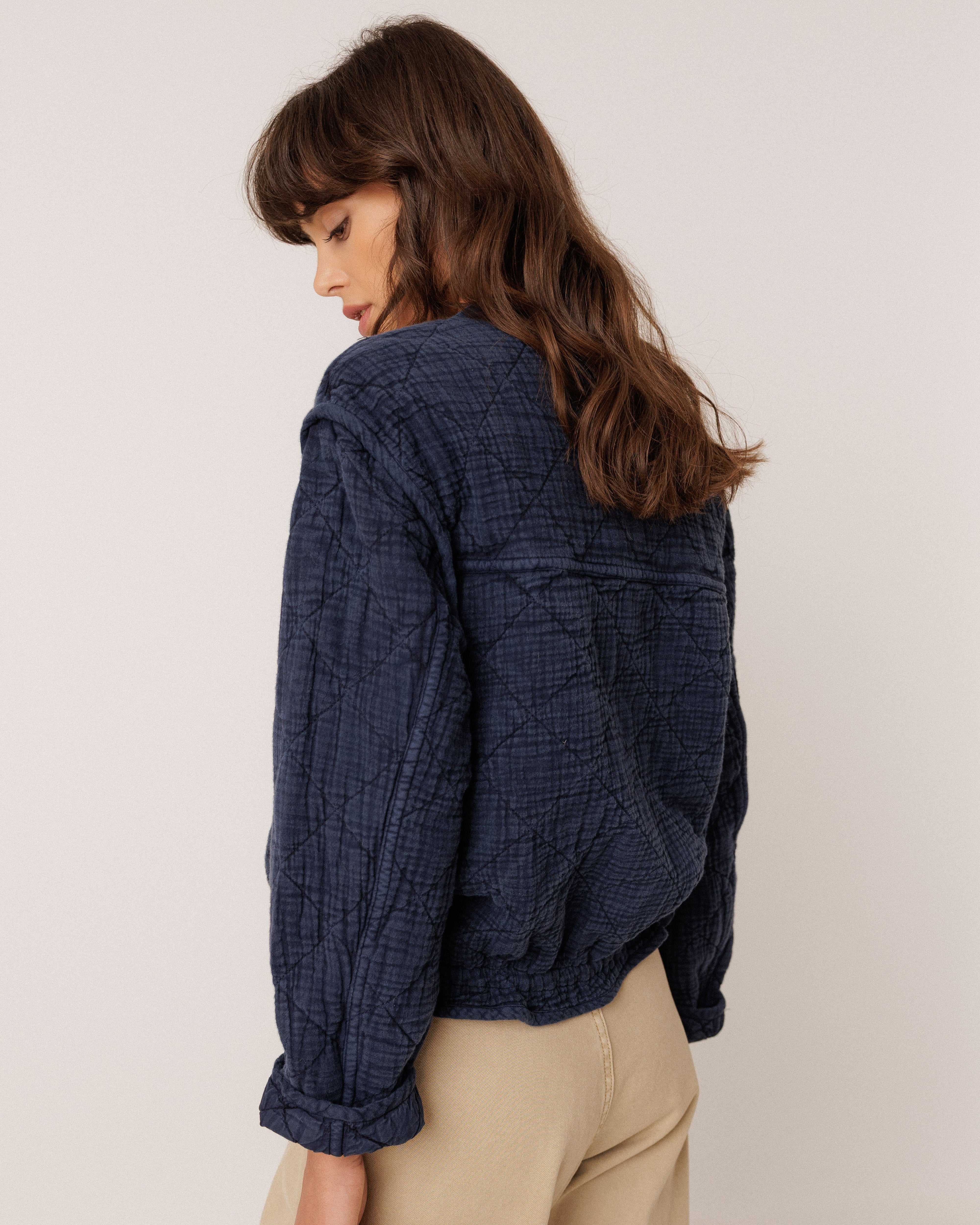 A woman with long brown hair is shown from a side-back angle wearing a dark navy crinkled textured jacket and beige trousers, photographed against a plain off-white studio background.