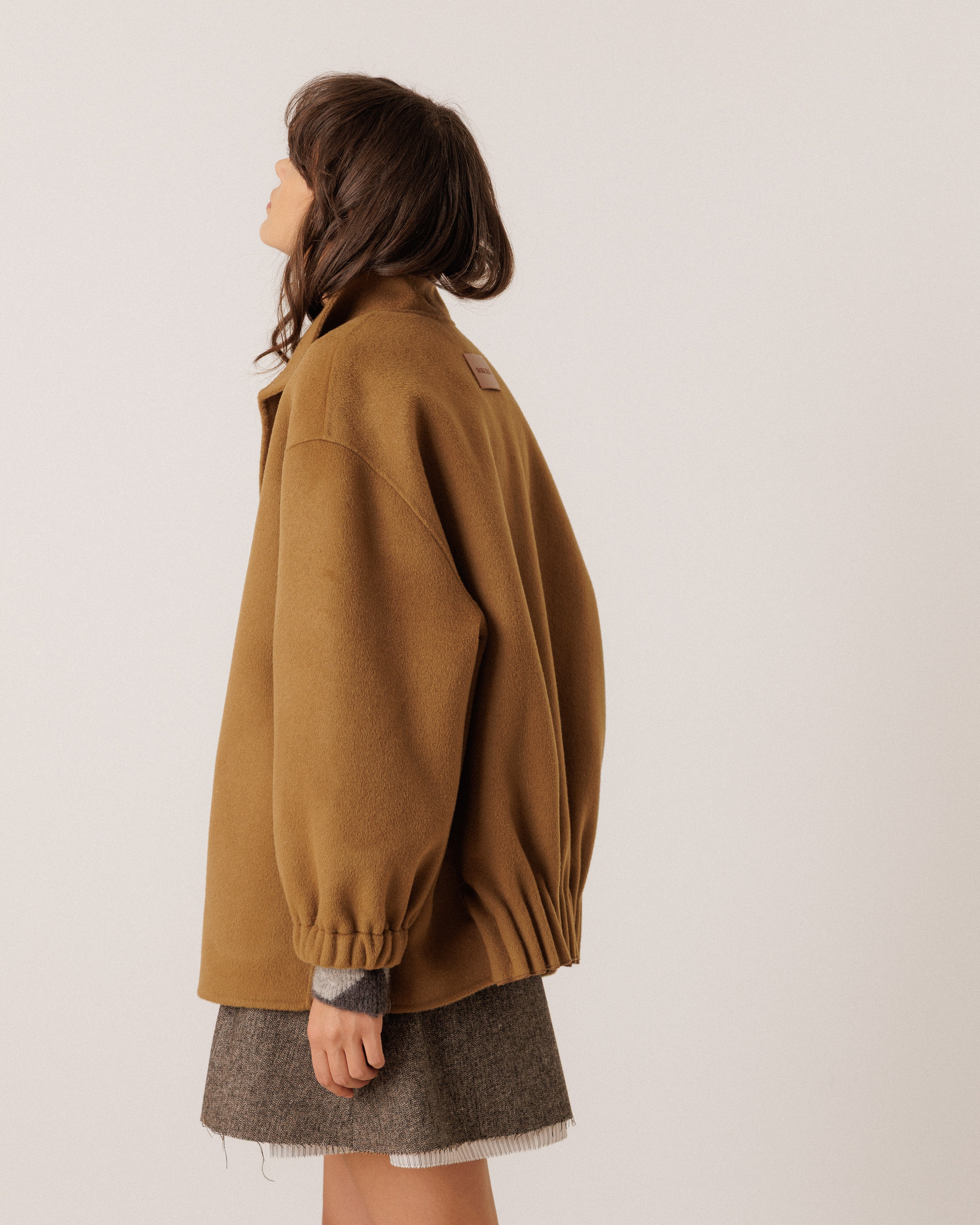 A woman shown from the side profile wearing an oversized camel brown jacket and a gray textured skirt, photographed against a plain light gray studio background.