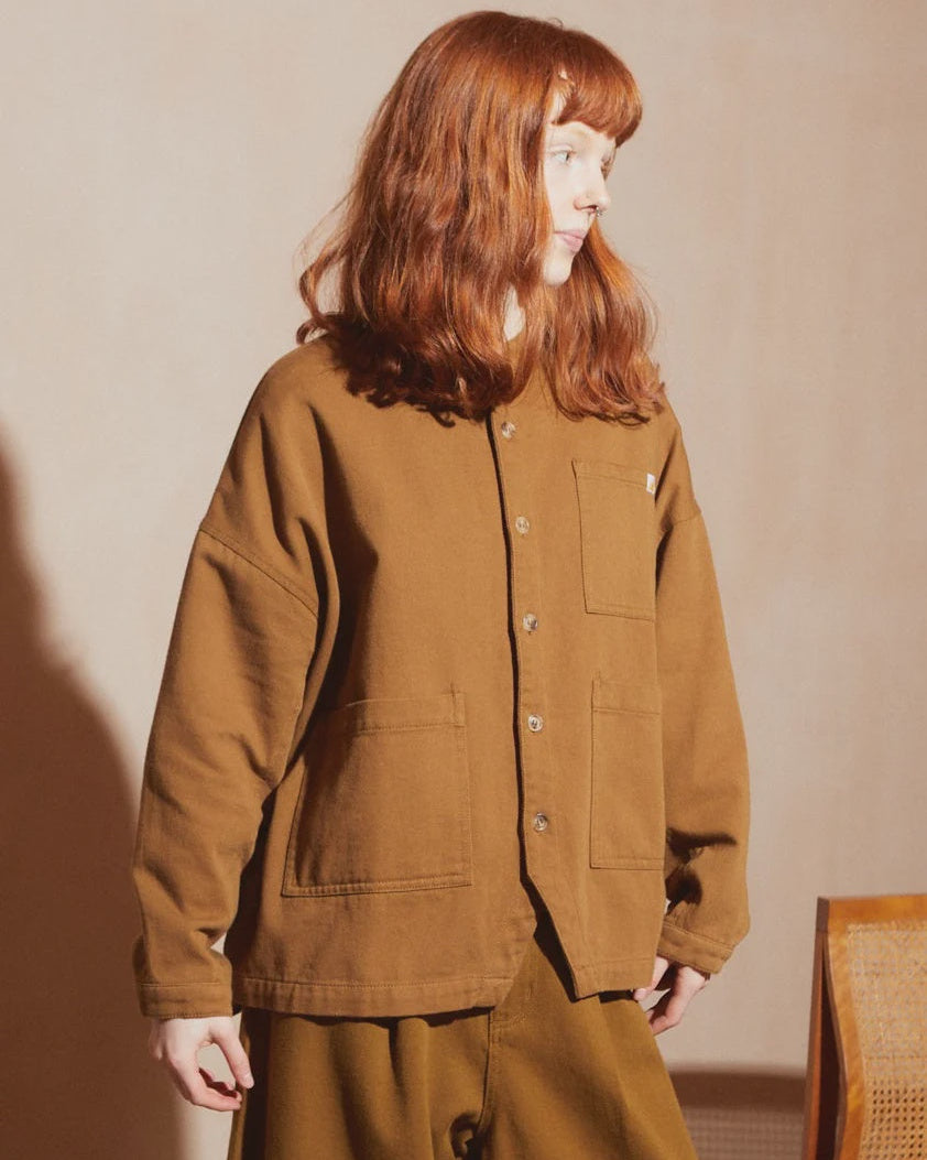 A woman with red hair wearing a brown button-up overshirt and matching brown pants, standing in a warm, sunlit studio setting.