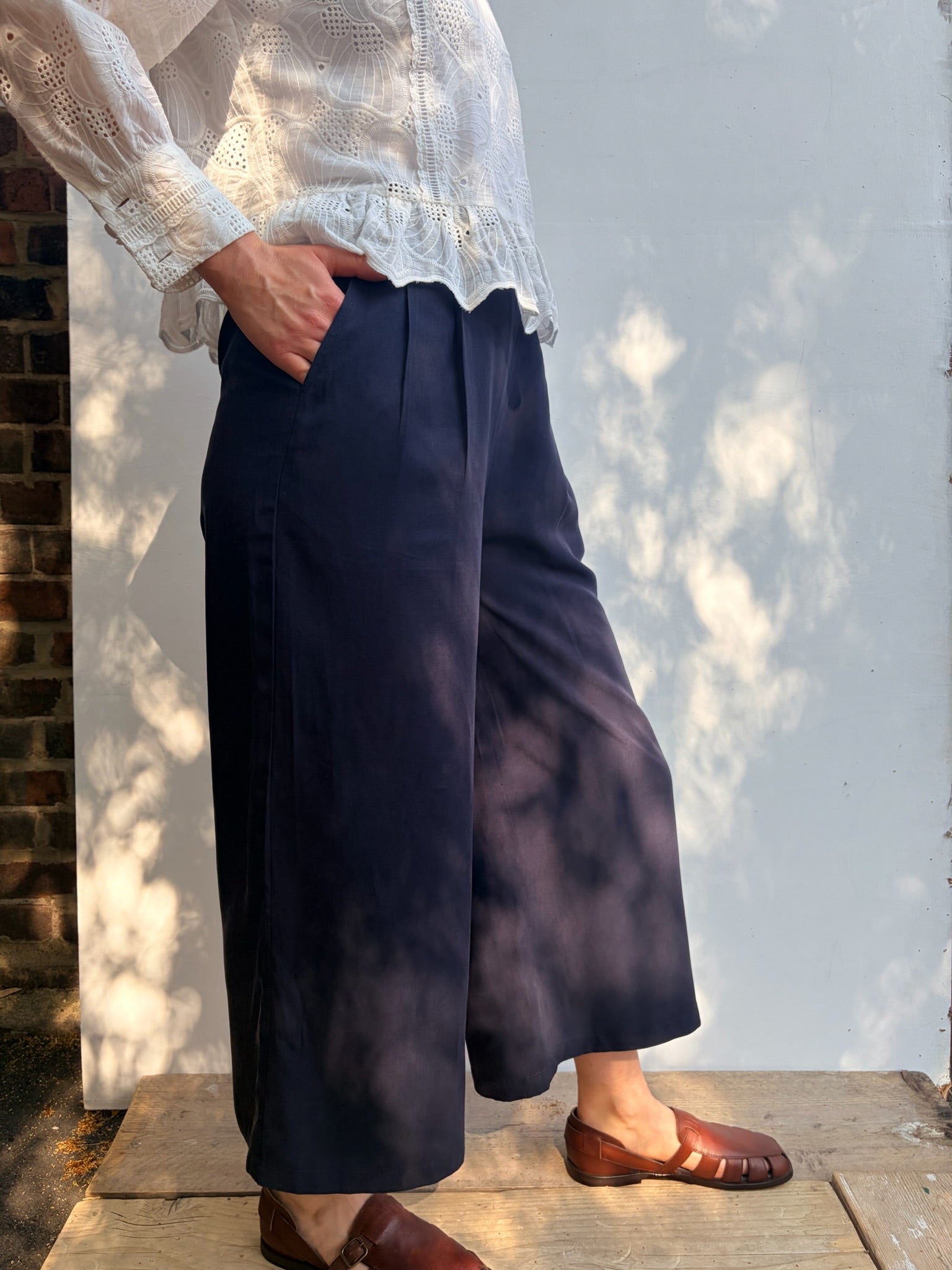 A person wearing navy wide-leg cropped trousers, a white lace-trim blouse, and brown leather loafers, standing outdoors against a white wall with dappled sunlight and shadows.
