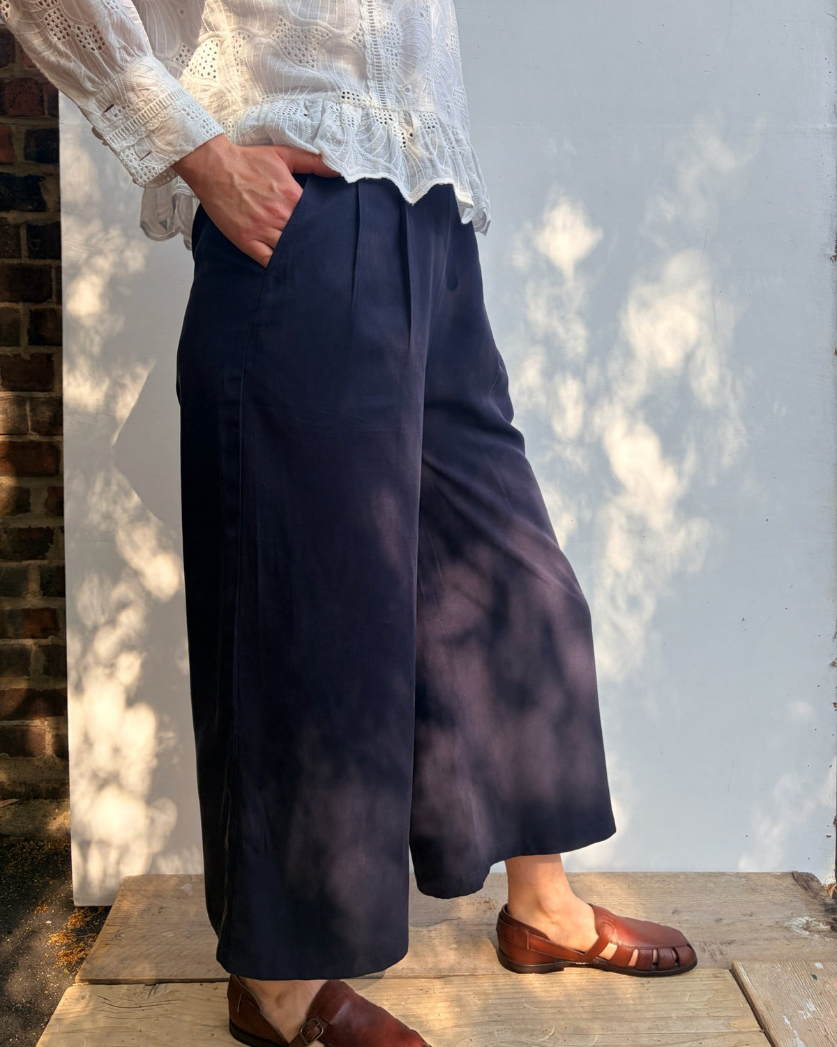A person wearing navy wide-leg cropped trousers, a white lace-trim blouse, and brown leather loafers, standing outdoors against a white wall with dappled sunlight and shadows.