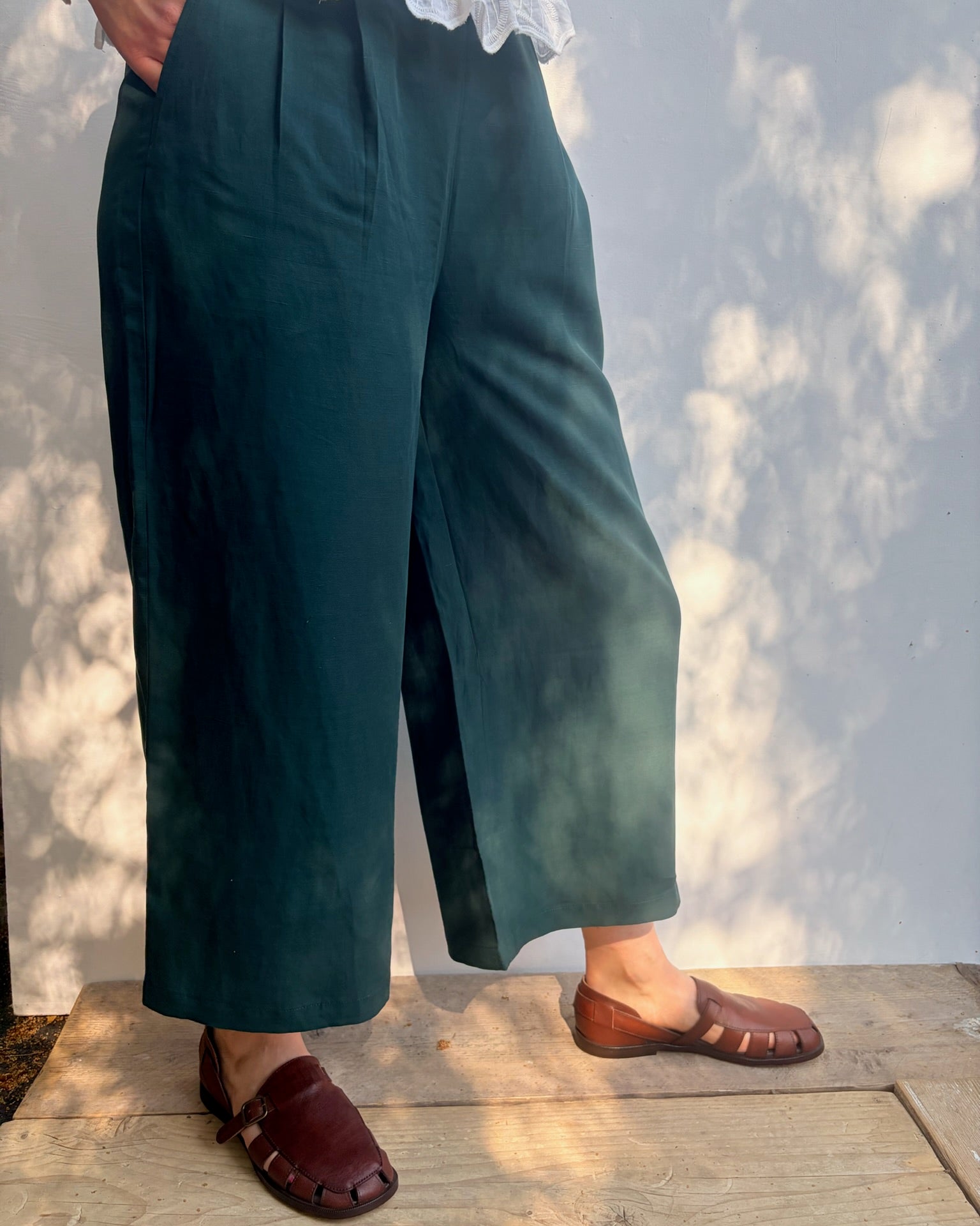 A person wearing dark teal wide-leg cropped pants with pleats, paired with brown leather slide sandals and a white lace-trim top, standing on a wooden deck against a wall with dappled sunlight.