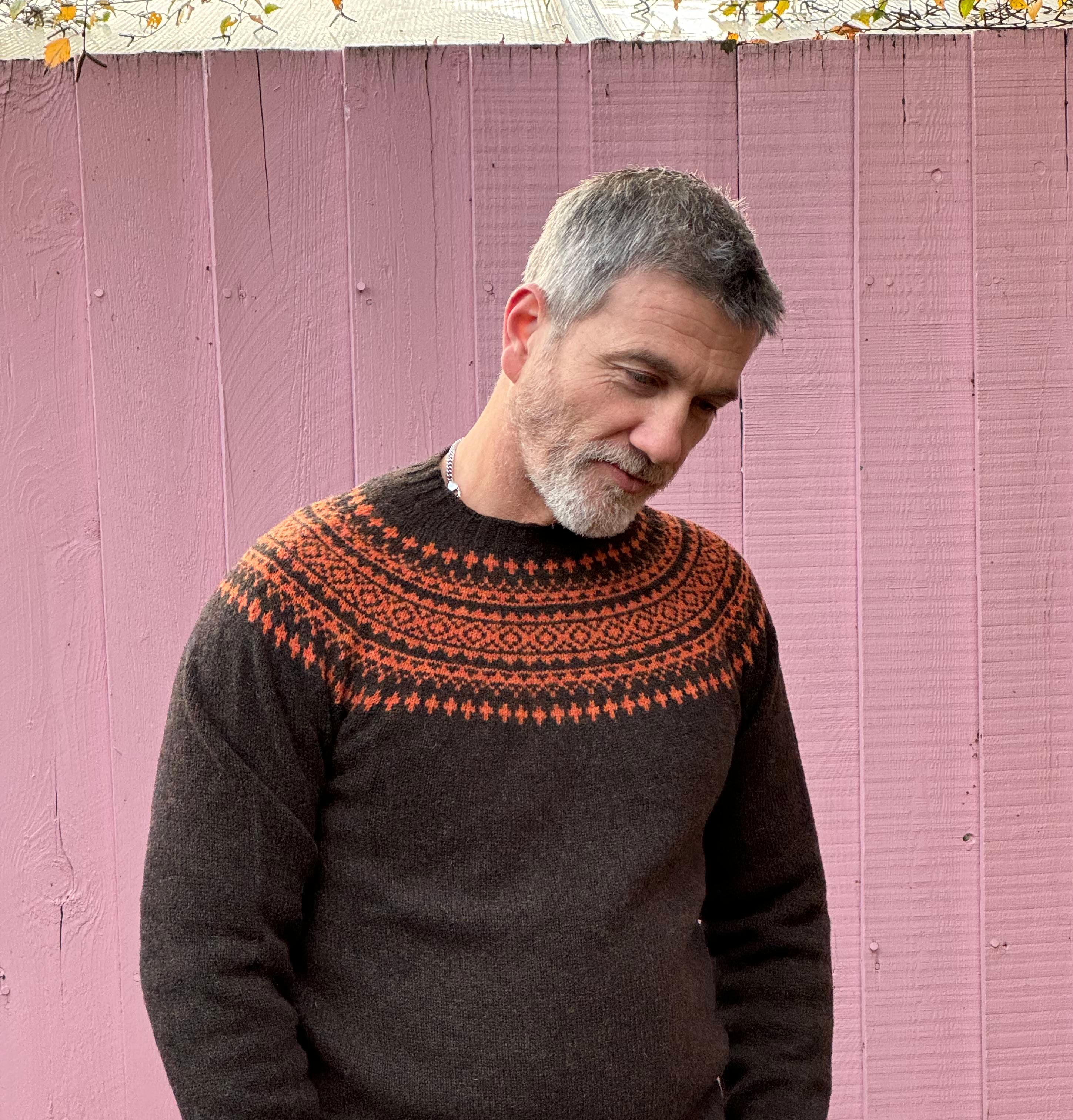 A man wearing a chocolate brown and orange fairisle pattern jumper with a ribbed round neck and long sleeves.