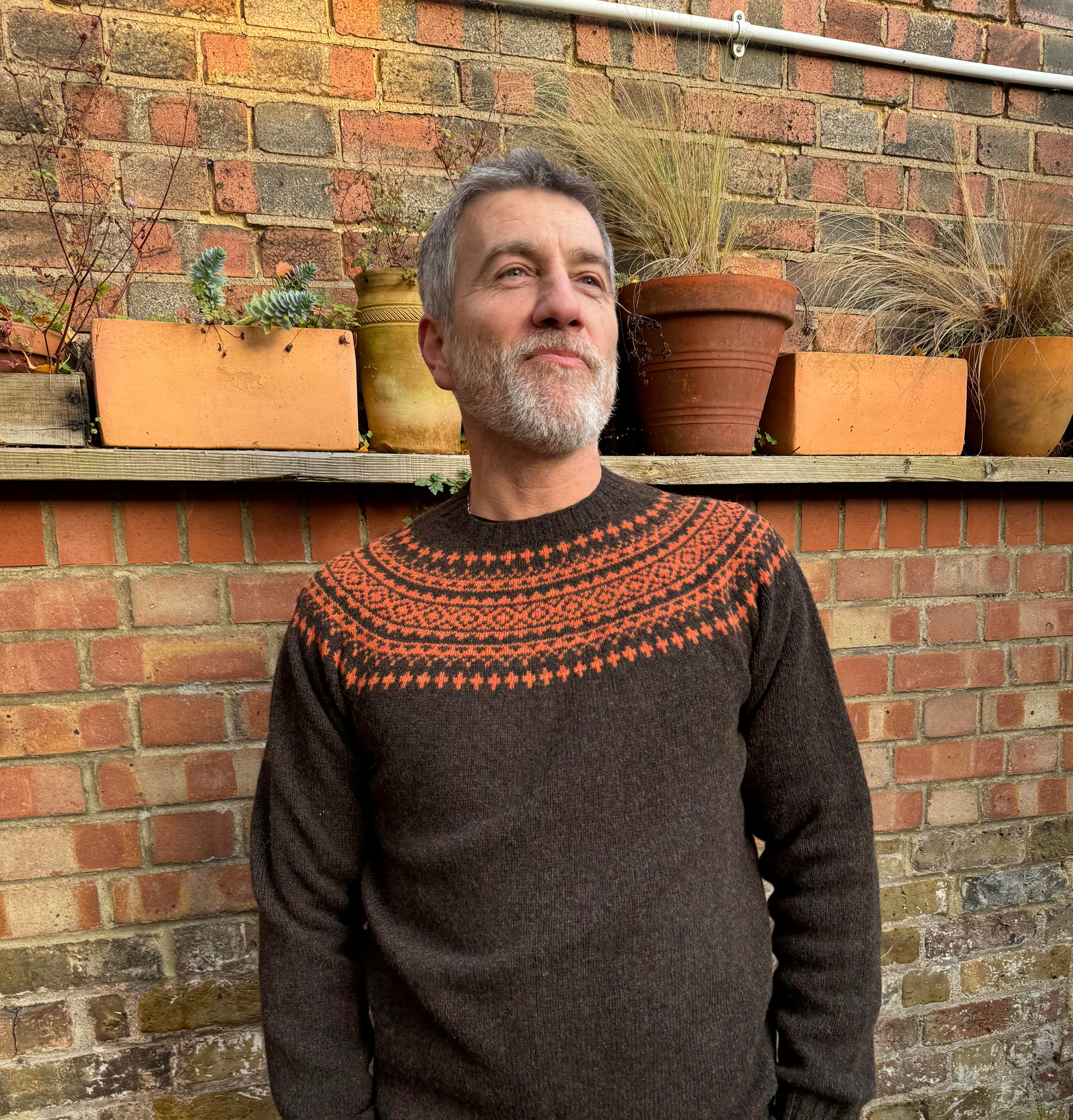 A man wearing a chocolate brown and orange fairisle pattern jumper with a ribbed round neck and long sleeves.