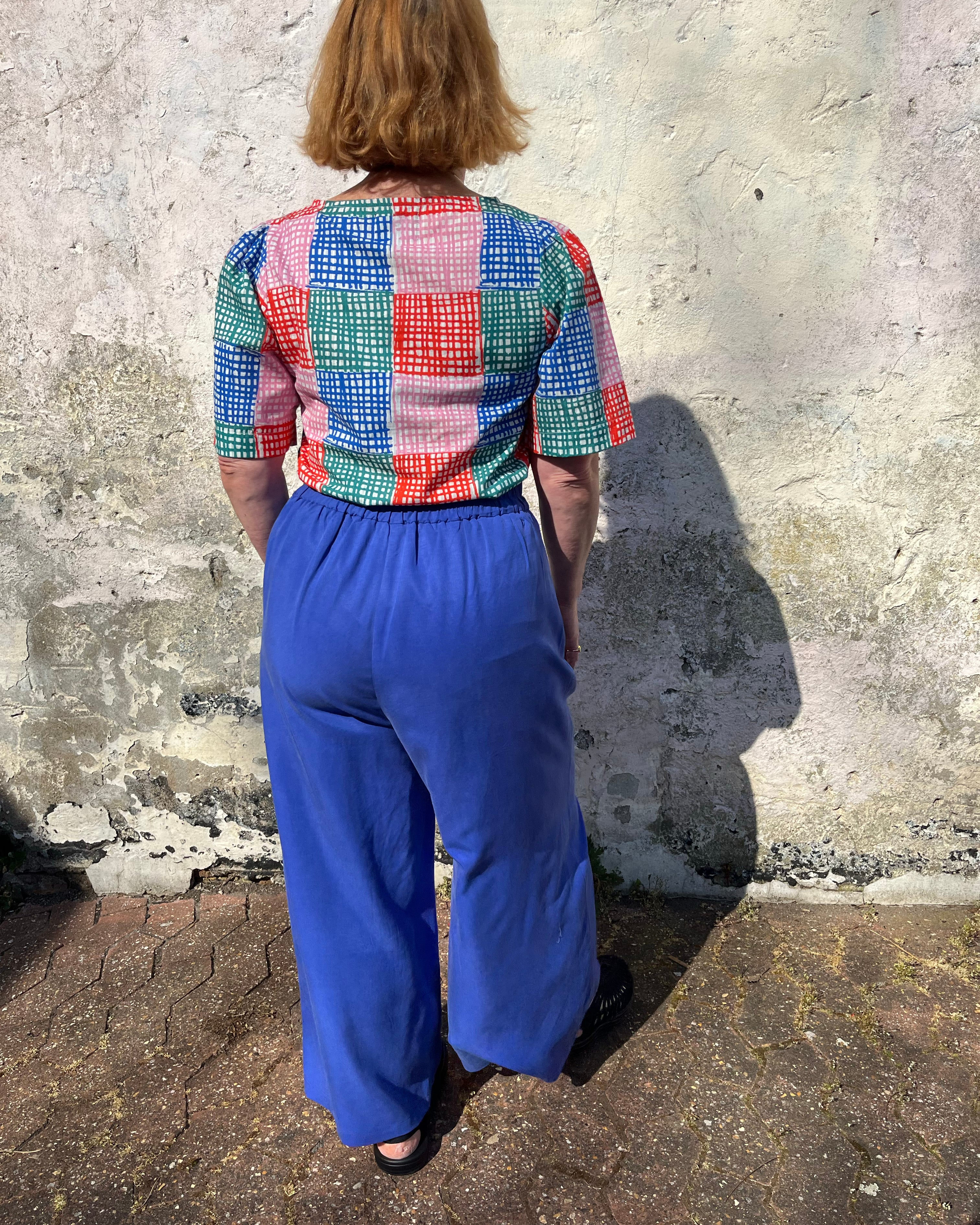 A person standing against a wall wearing cobalt lyocell wide-leg trousers with a flat front waistband, elasticated back, and pleats at the front.