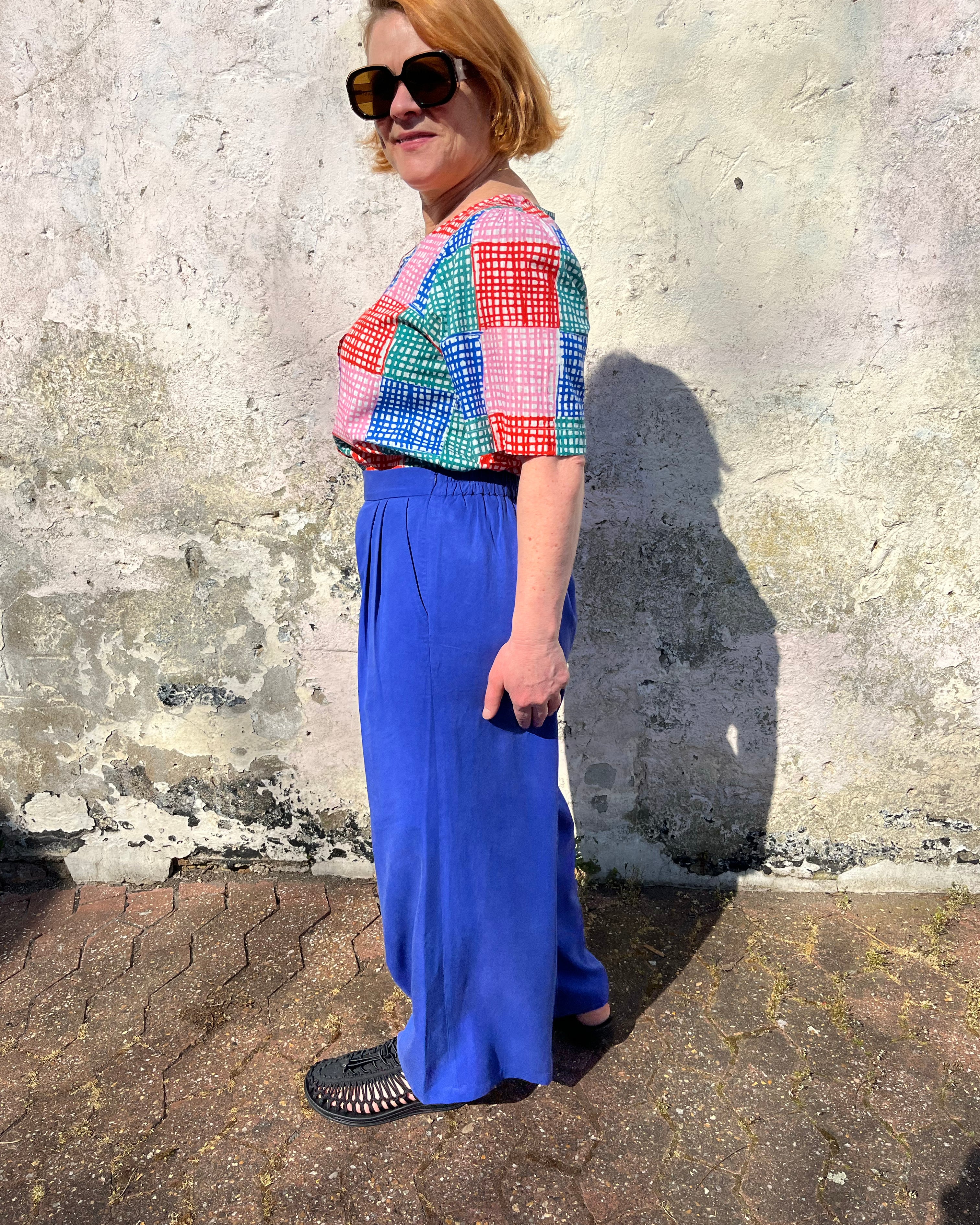 A person standing against a wall wearing cobalt lyocell wide-leg trousers with a flat front waistband, elasticated back, and pleats at the front.