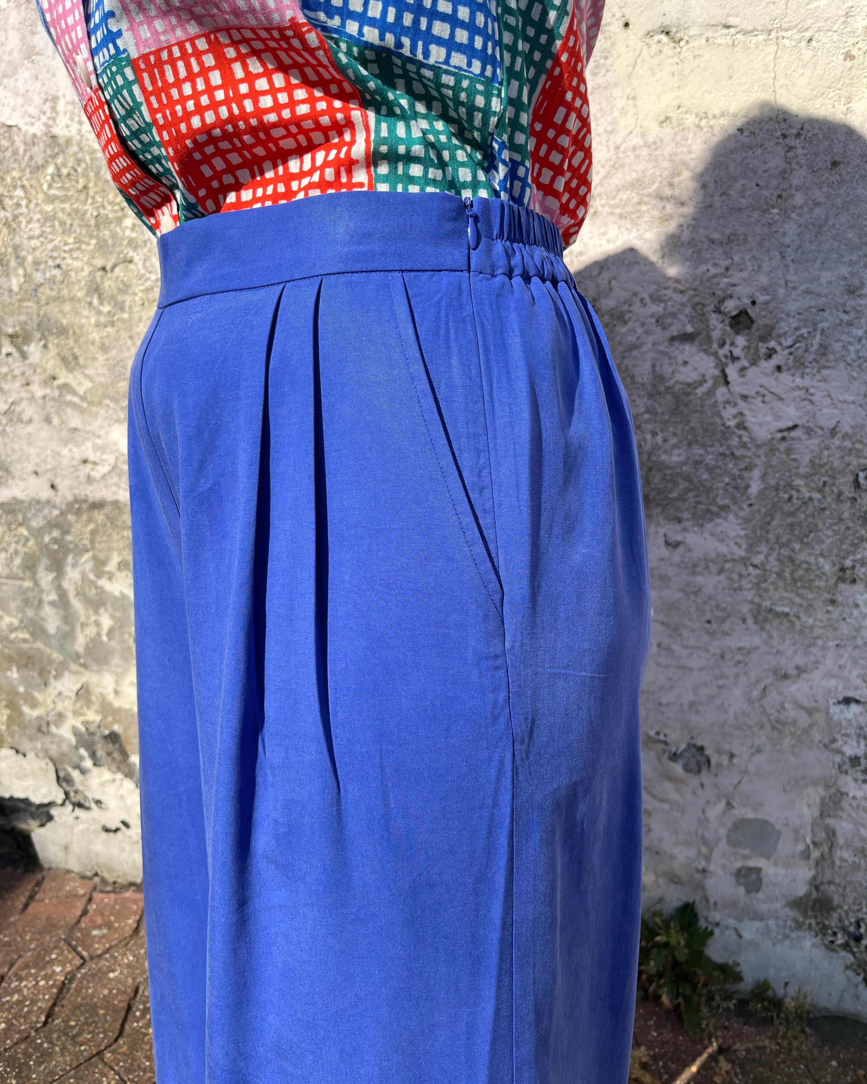 A person standing against a wall wearing cobalt lyocell wide-leg trousers with a flat front waistband, elasticated back, and pleats at the front.