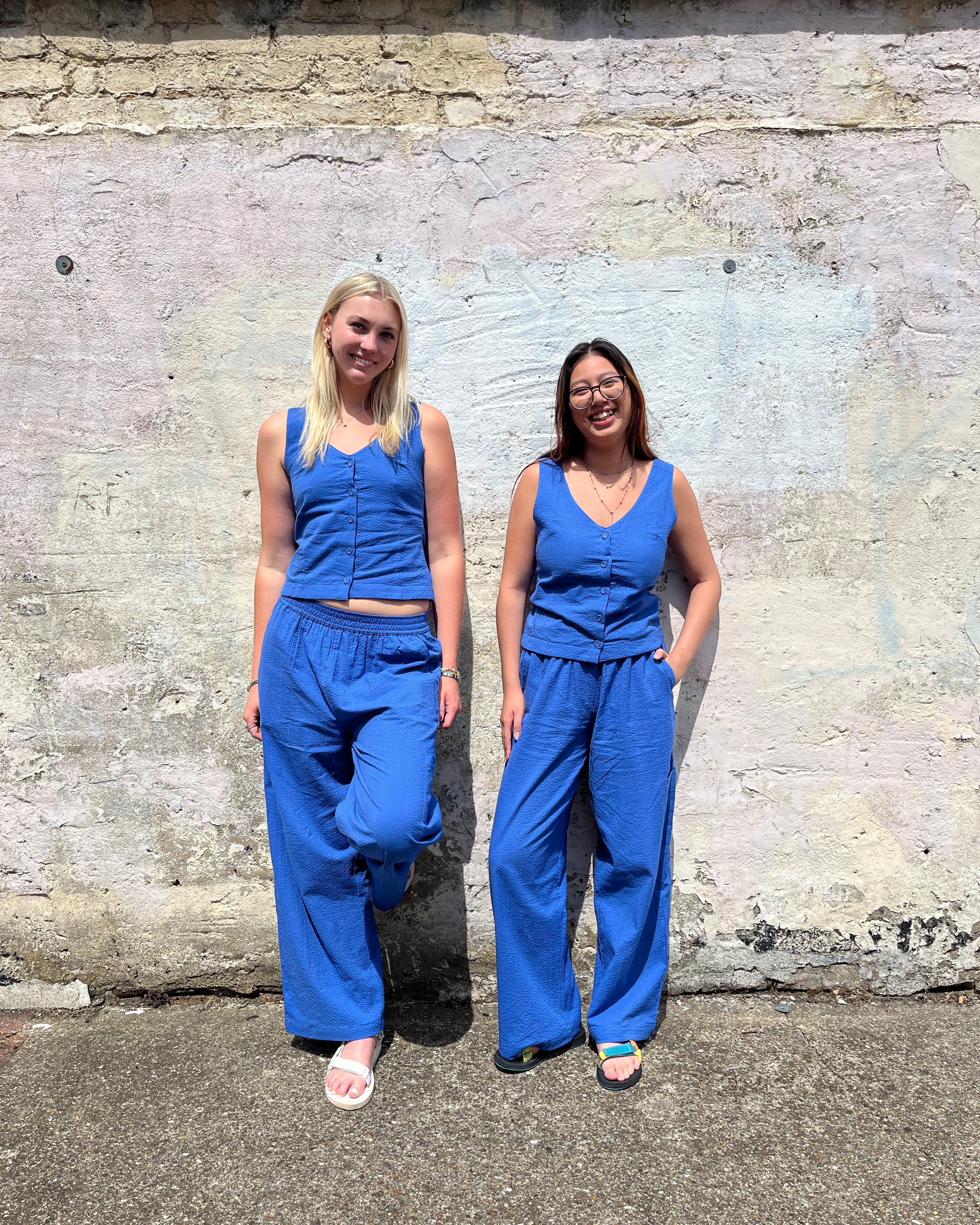 Two women wearing matching blue jumpsuits standing against a textured wall.