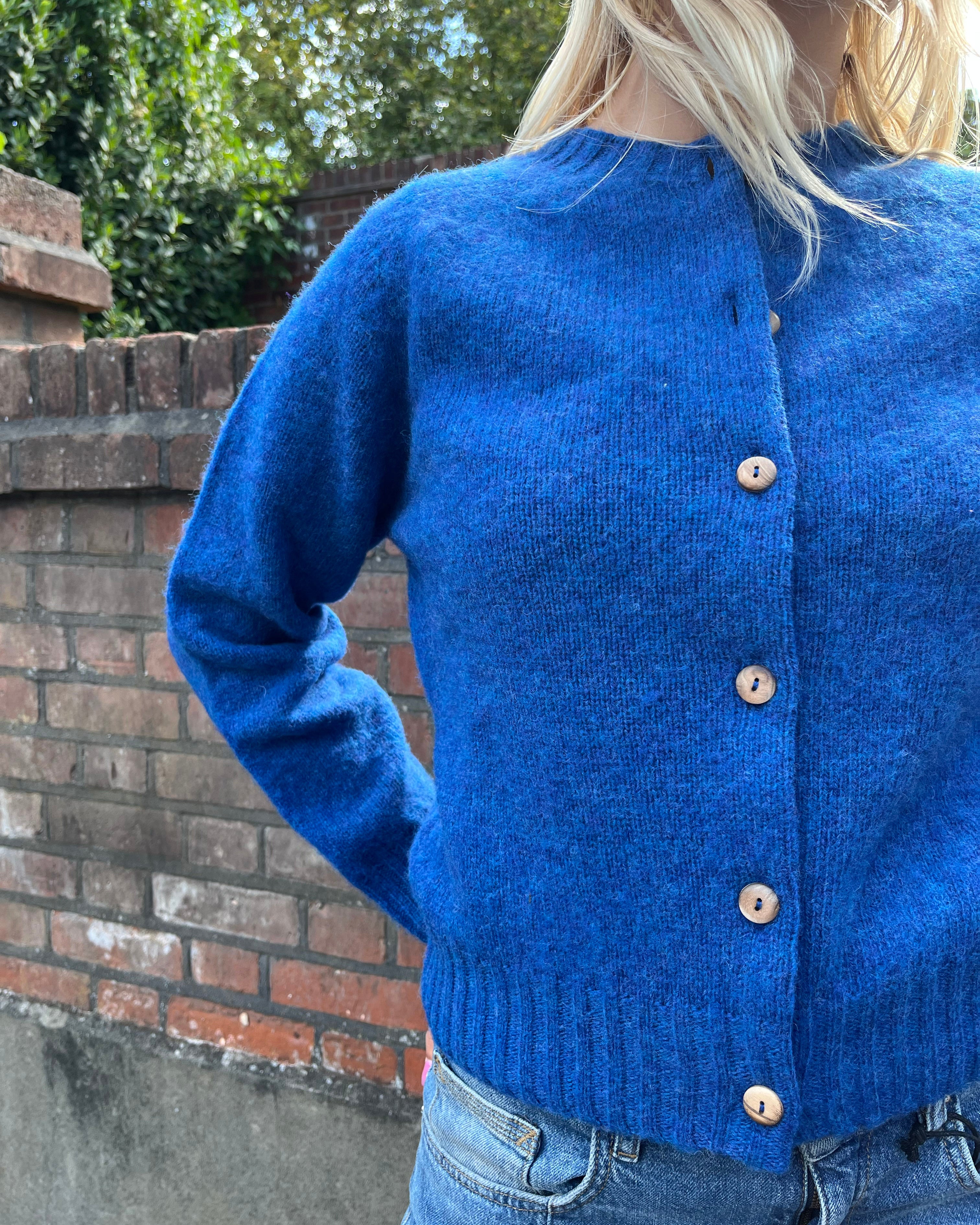 Person wearing a blue cardigan with a brick wall and greenery in the background