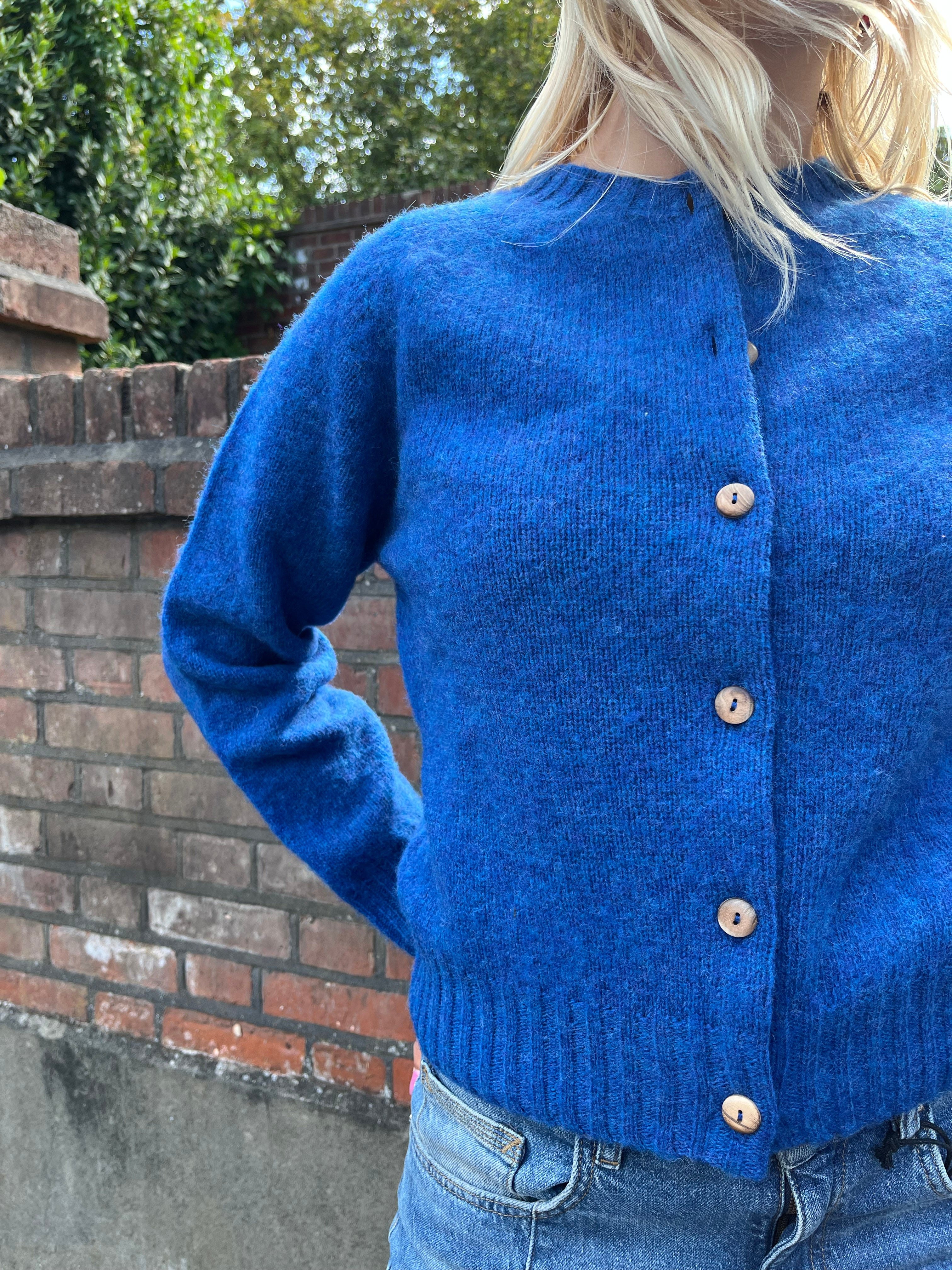 Person wearing a blue cardigan with a brick wall and greenery in the background