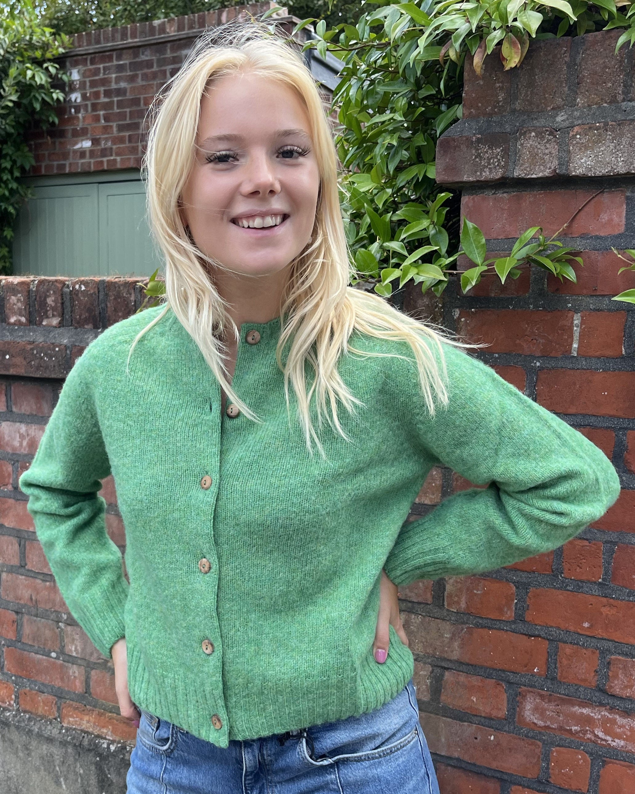 Person wearing a green cardigan standing in front of a brick wall with plants.