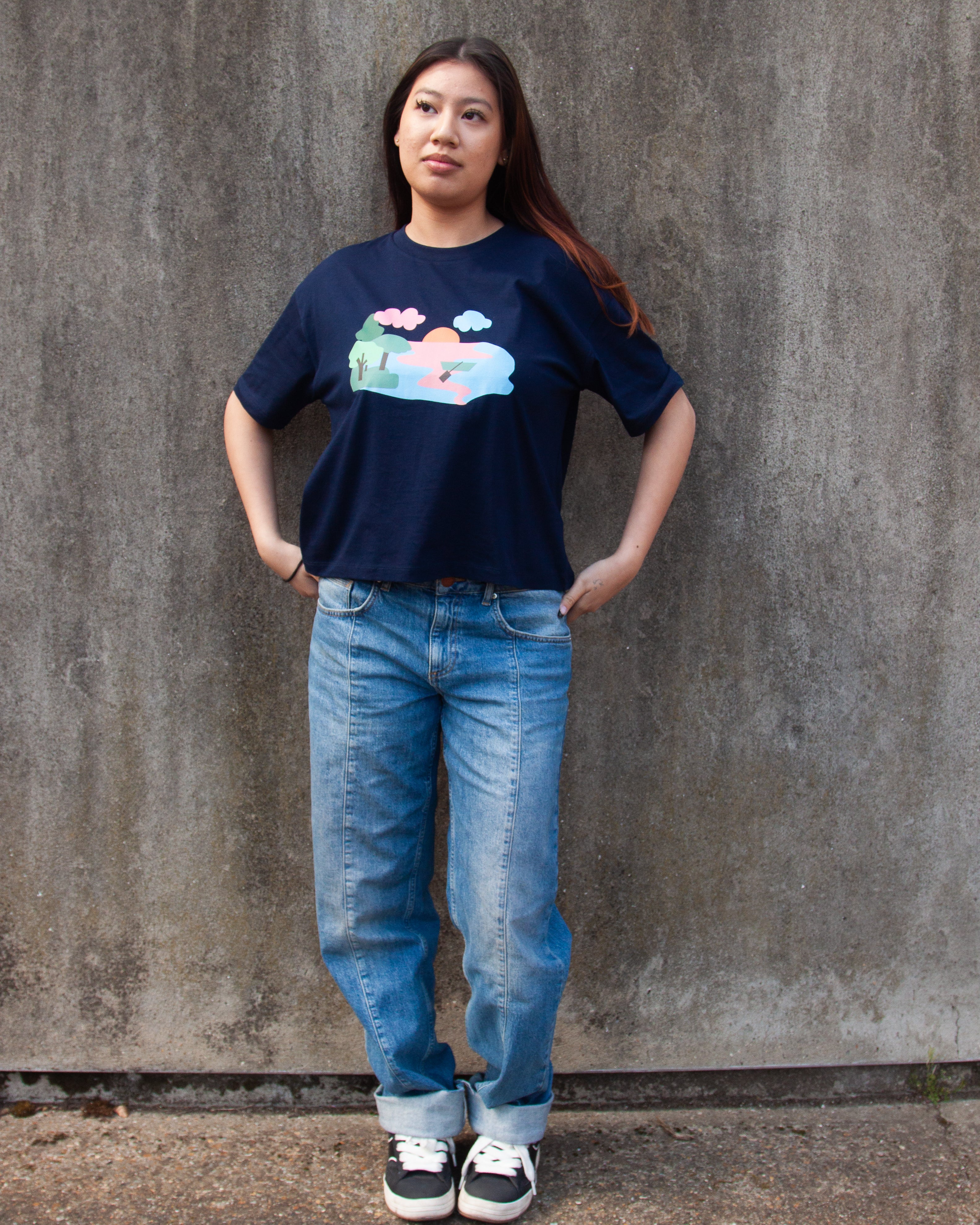A navy blue t-shirt with a graphic print of a lake scene at dusk with a lone canoe, pink and blue waters, and green trees.