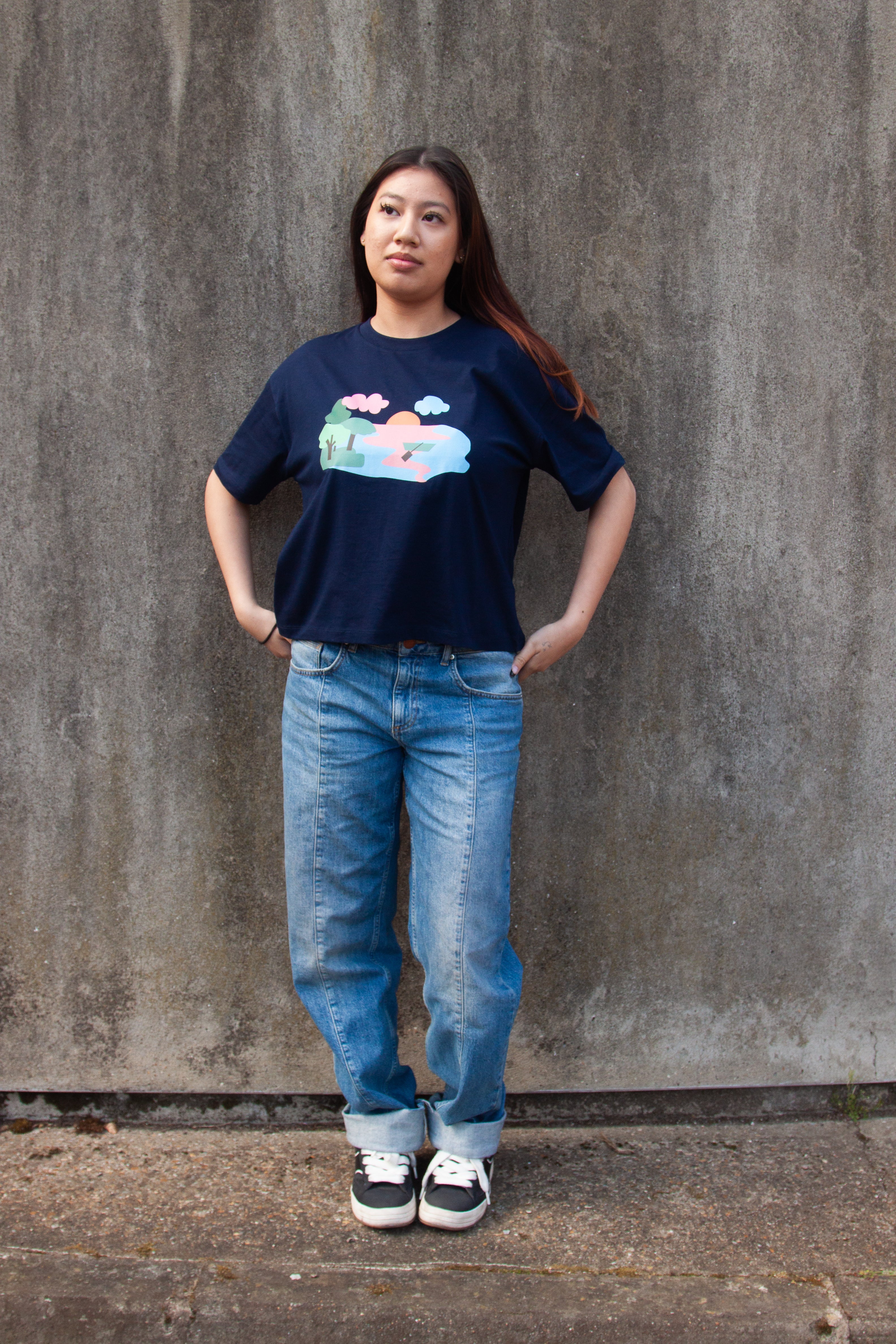 A navy blue t-shirt with a graphic print of a lake scene at dusk with a lone canoe, pink and blue waters, and green trees.