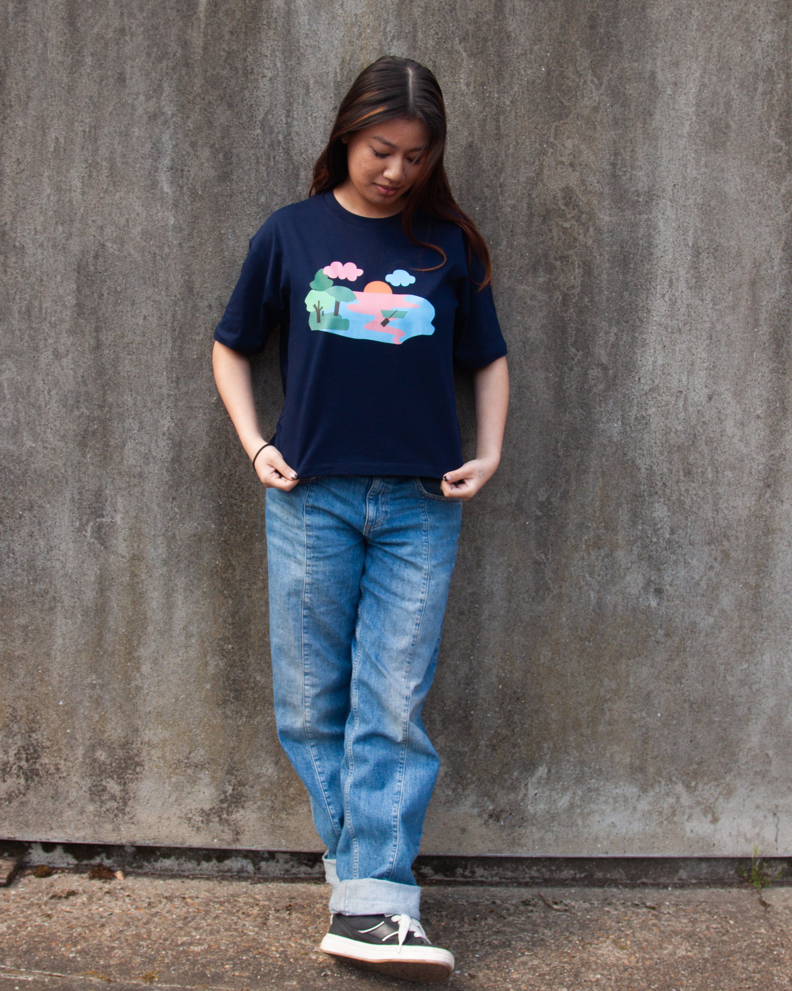 A navy blue t-shirt with a graphic print of a lake scene at dusk with a lone canoe, pink and blue waters, and green trees.