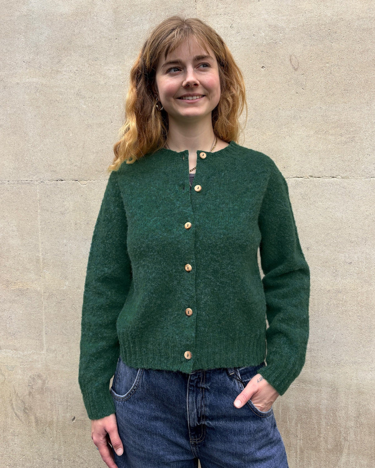A woman with shoulder-length light brown hair stands against a light beige stone wall, wearing a dark green knit button-front cardigan with wooden buttons and blue jeans.