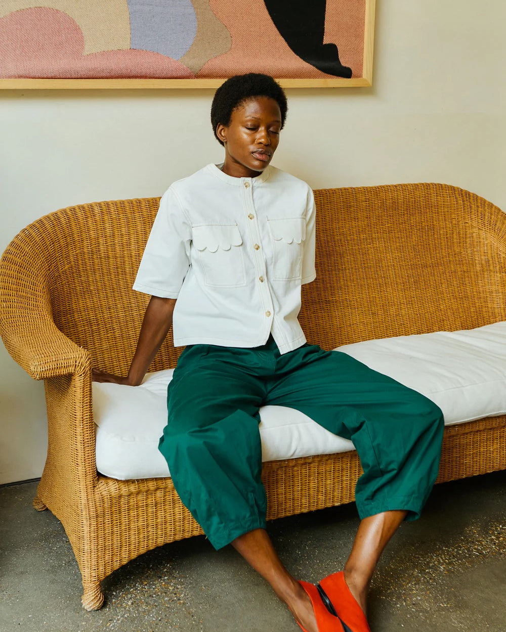 Woman sitting on a wicker couch wearing a white jacket and green pants, with a colorful abstract painting in the background.