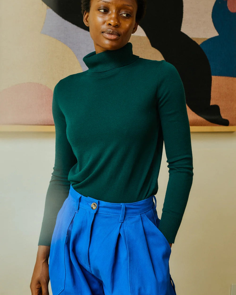 Person wearing a green turtleneck and blue pants against a colorful abstract wall.