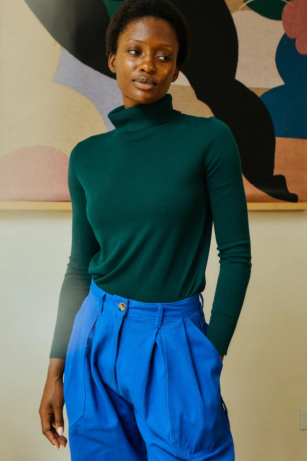 Person wearing a green turtleneck and blue pants against a colorful abstract wall.