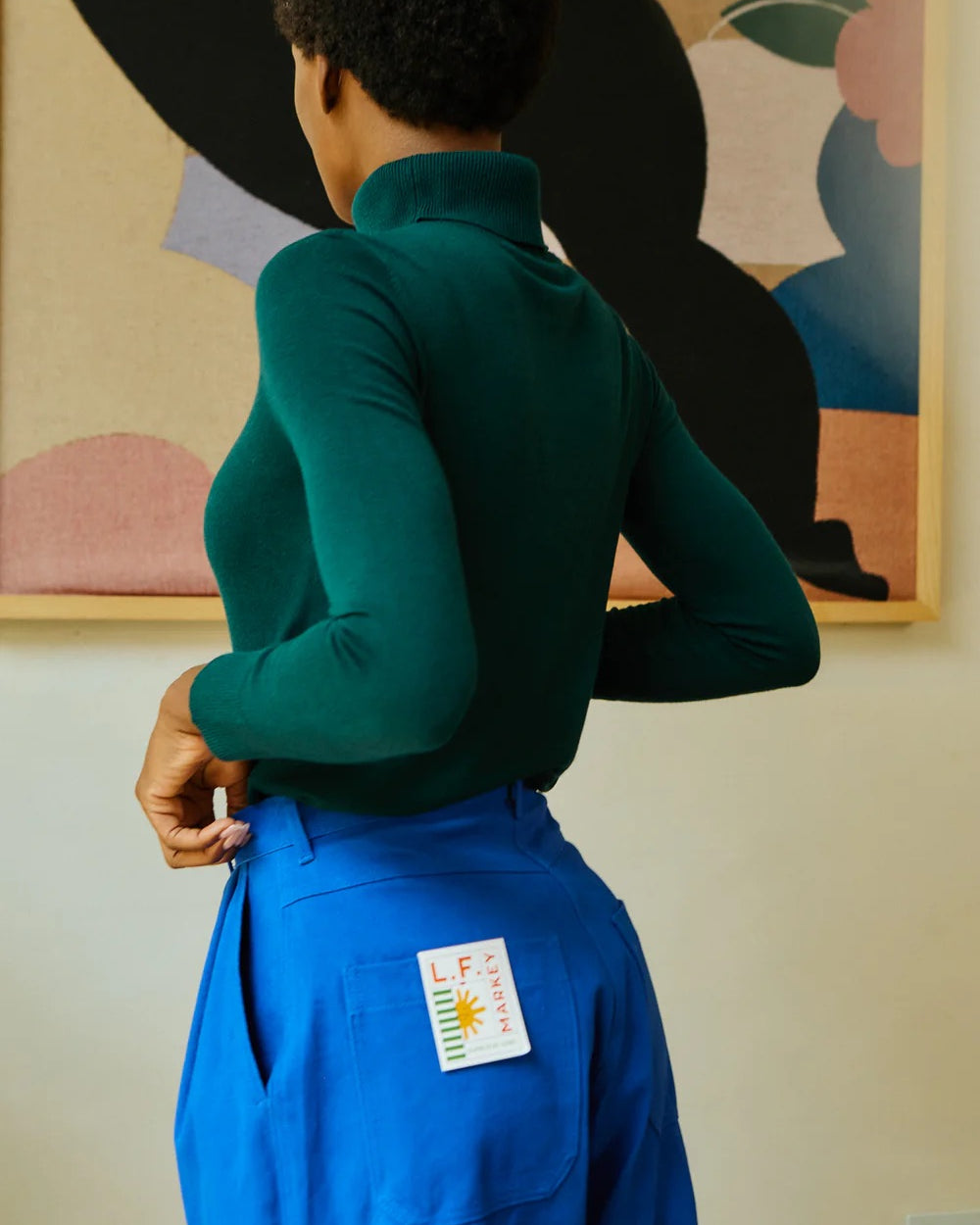 Person wearing a green sweater and blue pants with a colorful abstract painting in the background.