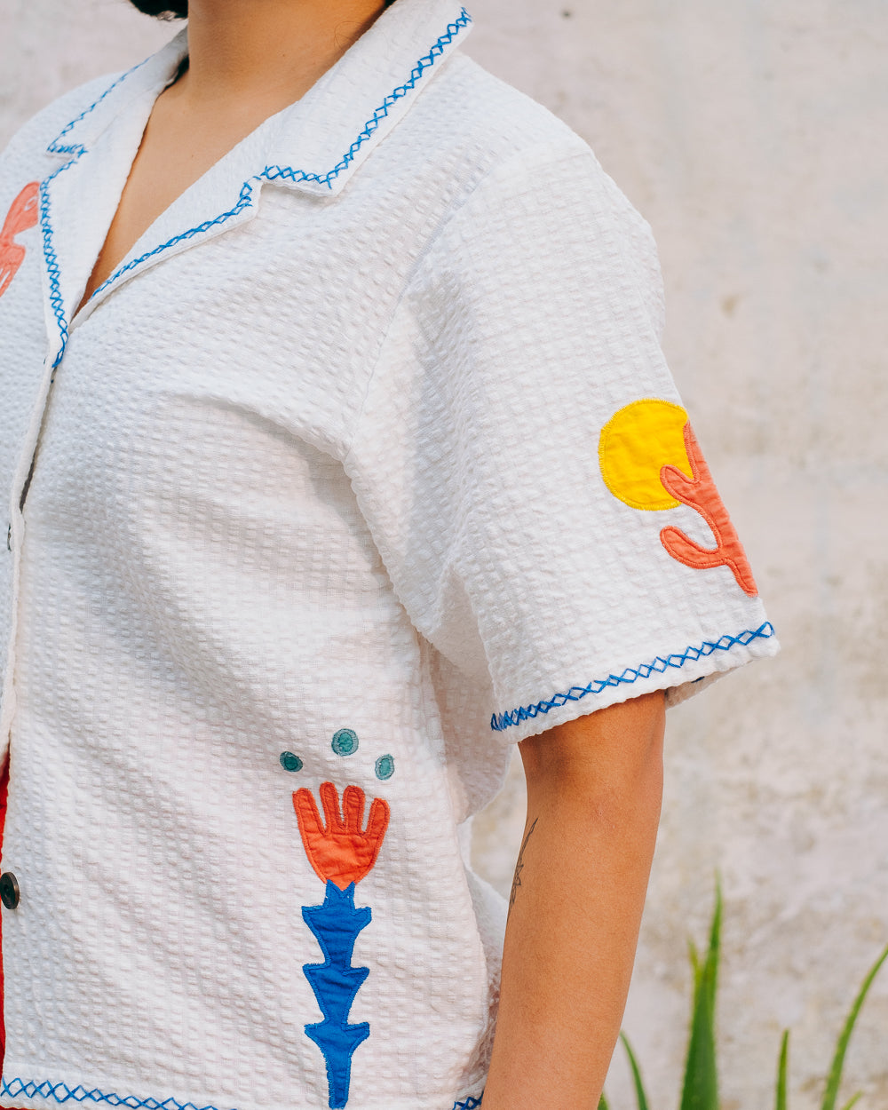 A white cotton blouse with colorful hand embroidery details, featuring a notched collar, wide cut sleeves, and a boxy fit.