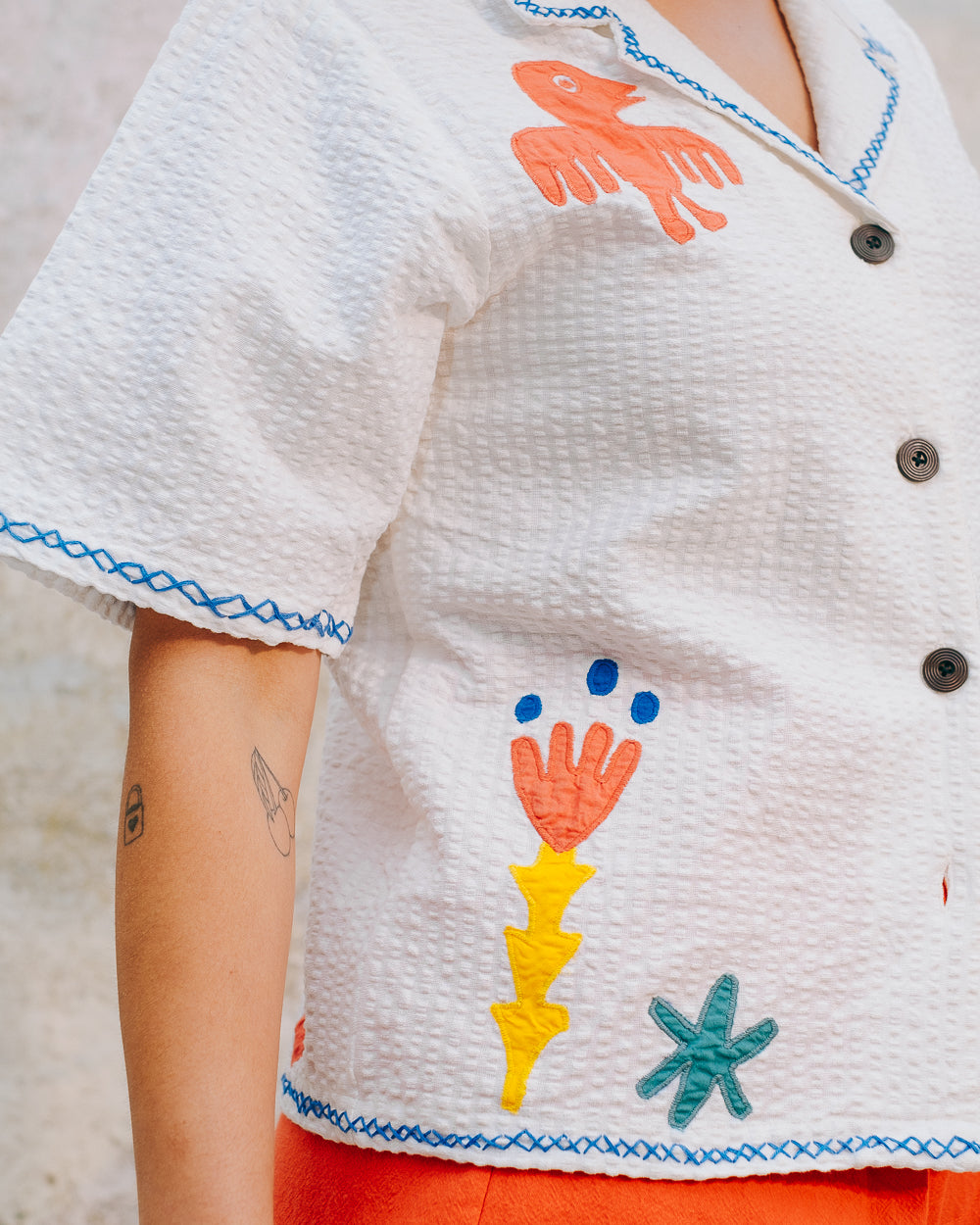 A white cotton blouse with colorful hand embroidery details, featuring a notched collar, wide cut sleeves, and a boxy fit.