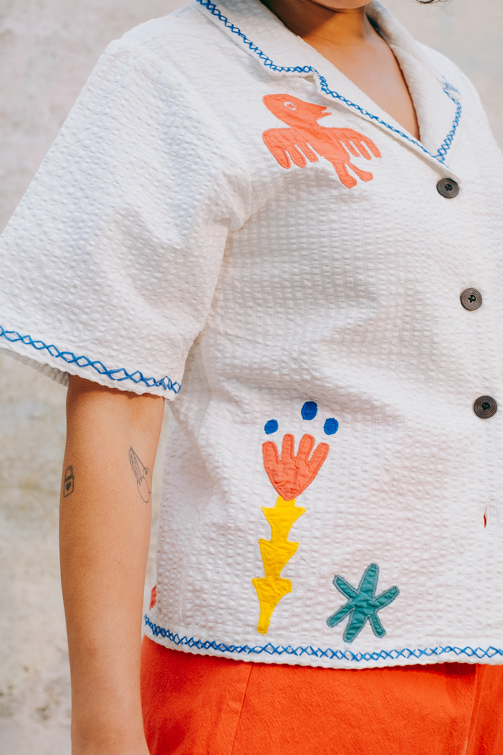 A white cotton blouse with colorful hand embroidery details, featuring a notched collar, wide cut sleeves, and a boxy fit.