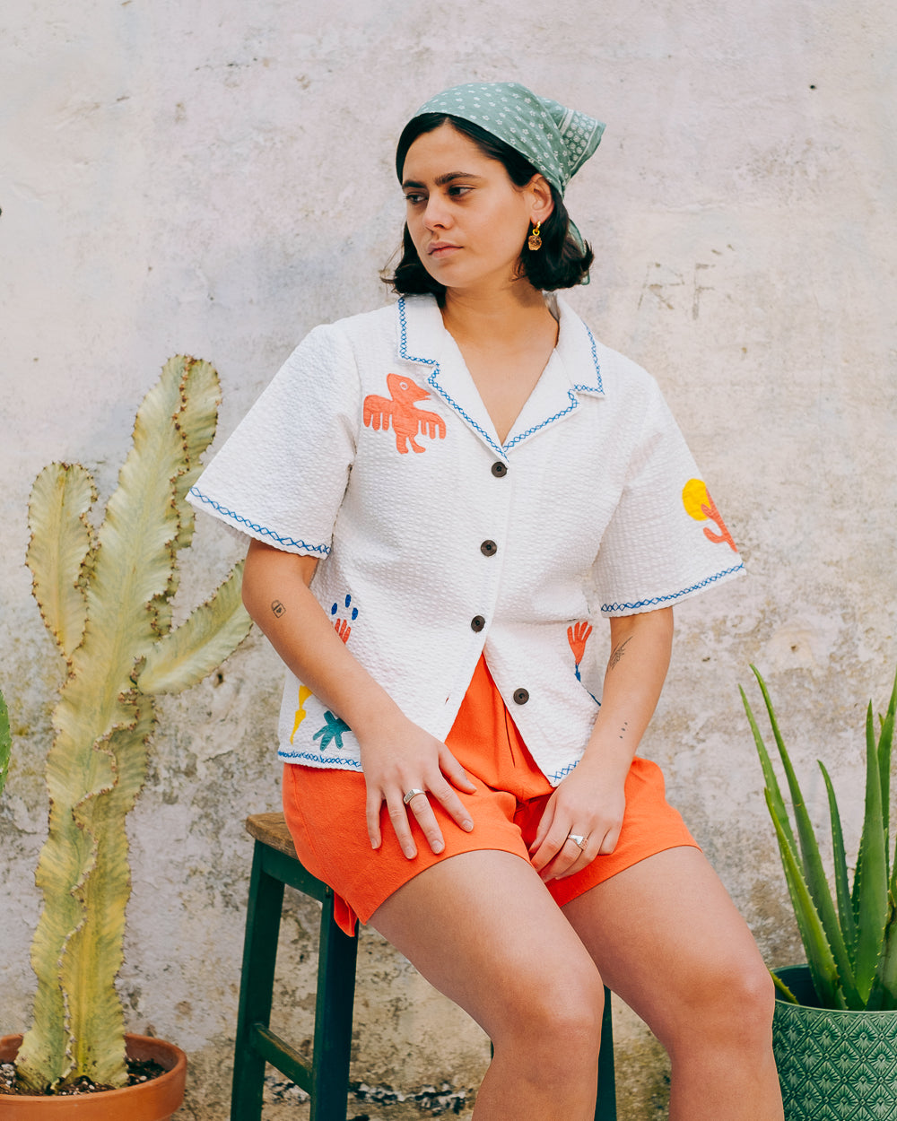 A white cotton blouse with colorful hand embroidery details, featuring a notched collar, wide cut sleeves, and a boxy fit.