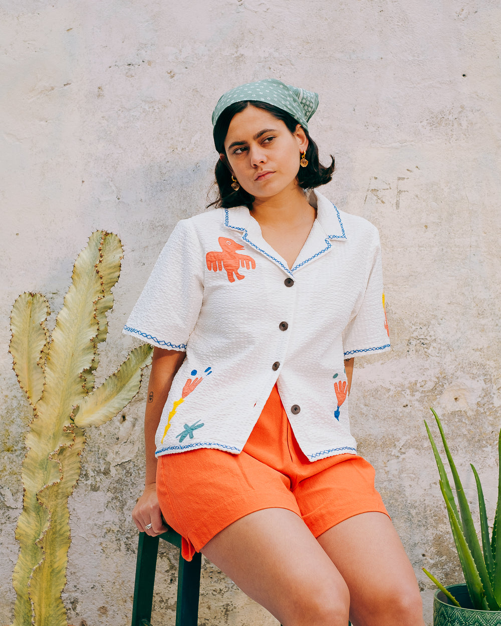 A white cotton blouse with colorful hand embroidery details, featuring a notched collar, wide cut sleeves, and a boxy fit.
