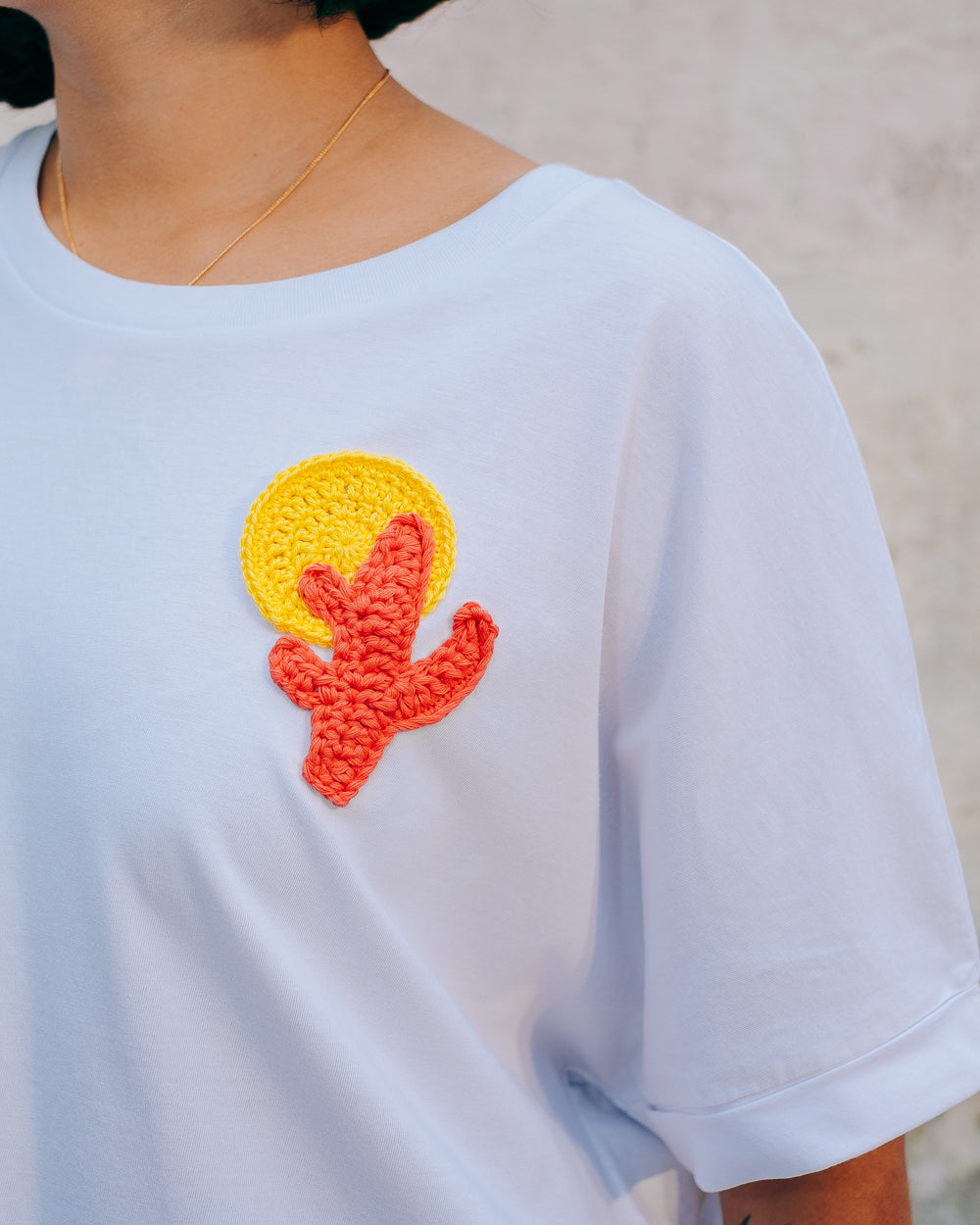 A person wearing a white crew neck t-shirt with a hand-crocheted red and yellow cactus and sun motif, paired with a blue skirt and a red belt.
