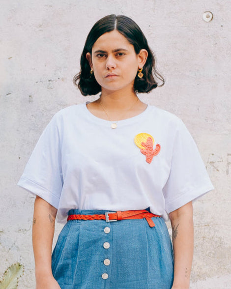 A person wearing a white crew neck t-shirt with a hand-crocheted cactus and sun design, paired with a blue skirt and an orange belt.